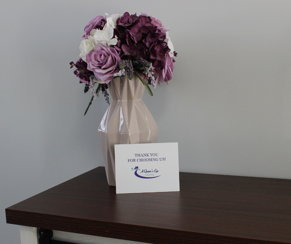 A bouquet of purple, white, and dark red flowers in a white ceramic vase on a dark brown table with a thank you card that reads, "Thank you for choosing us!" from A Queen's Eye Cleaning.