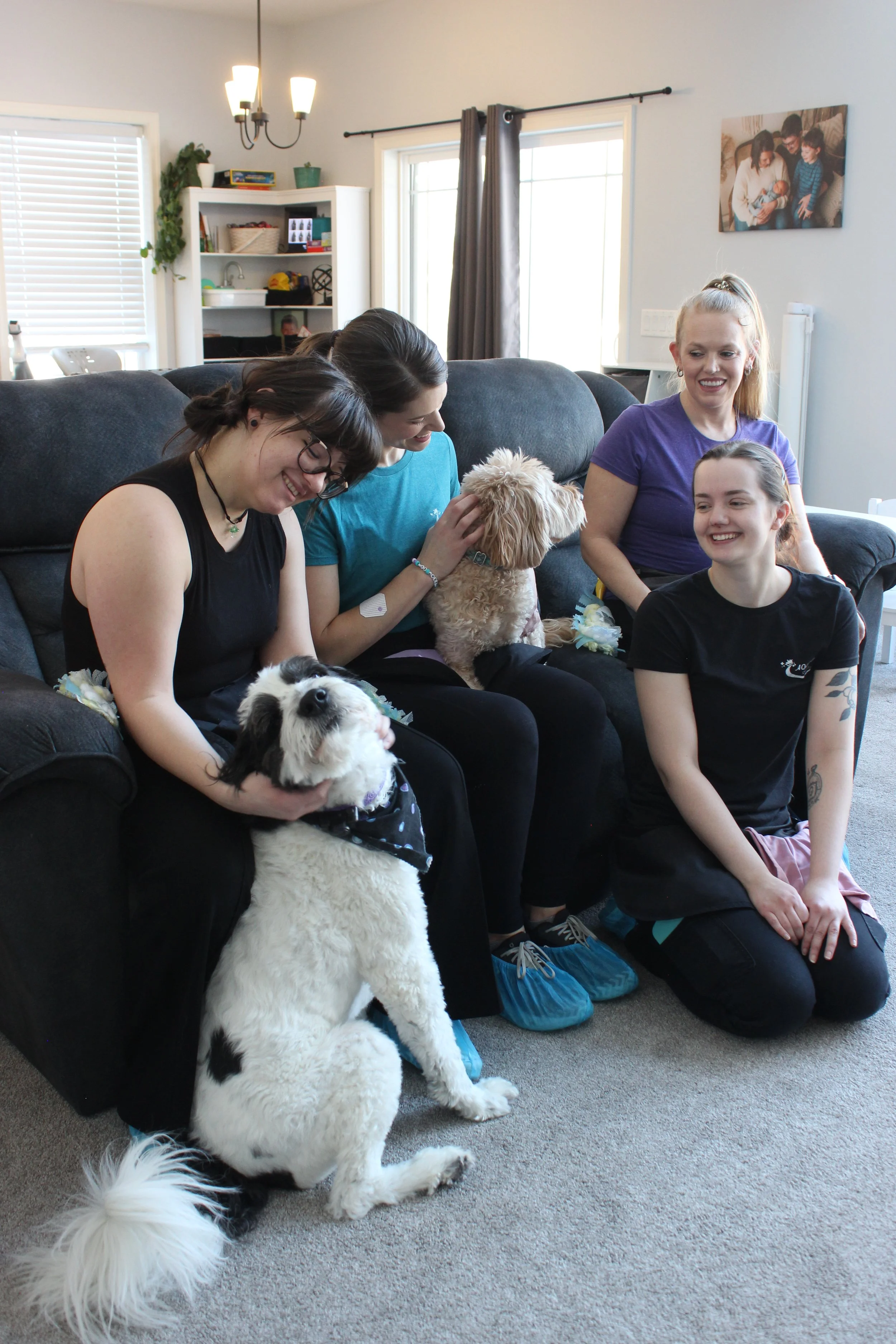 Four women and three dogs sitting on a couch and on the floor, smiling and enjoying each other's company in a living room.
