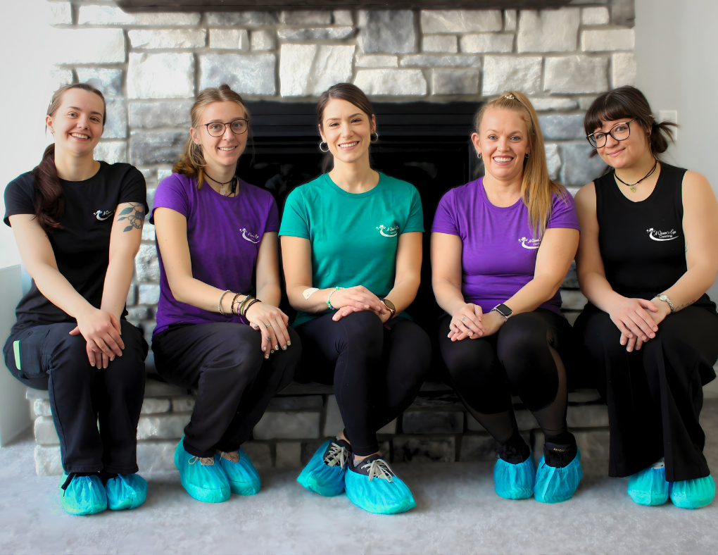 Five young women sitting on a black bench in front of a stone fireplace, smiling at the camera. They are wearing colorful t-shirts and shoe covers. The women are dressed casually, with some wearing glasses and jewelry.