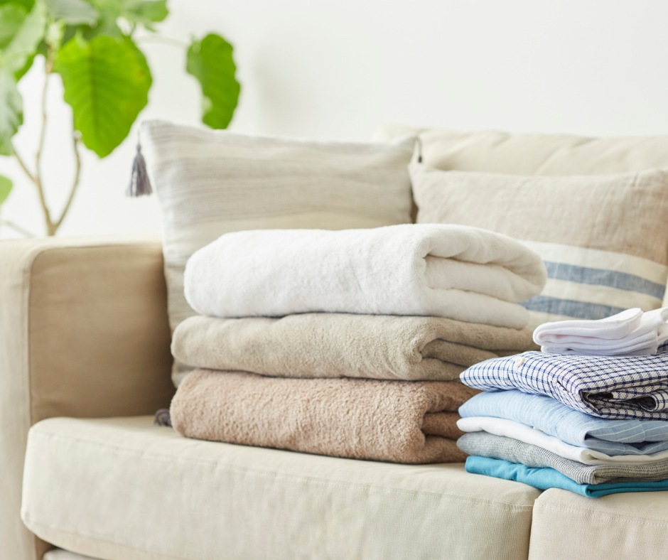 Folded towels and clothing on a light-colored sofa with pillows, in a living room with a potted plant in the background.