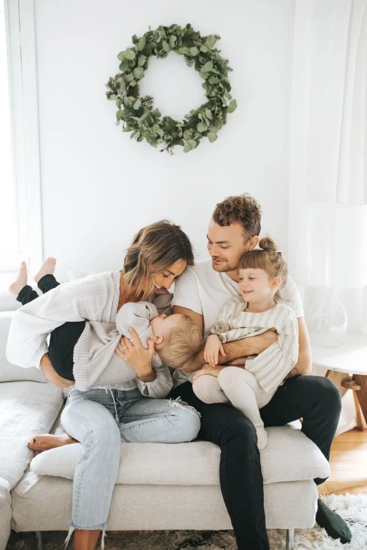 Family of four sitting on a couch in a bright living room, with a green wreath hanging on the white wall above them. The mother, father, young girl, and baby are smiling and enjoying their time together.