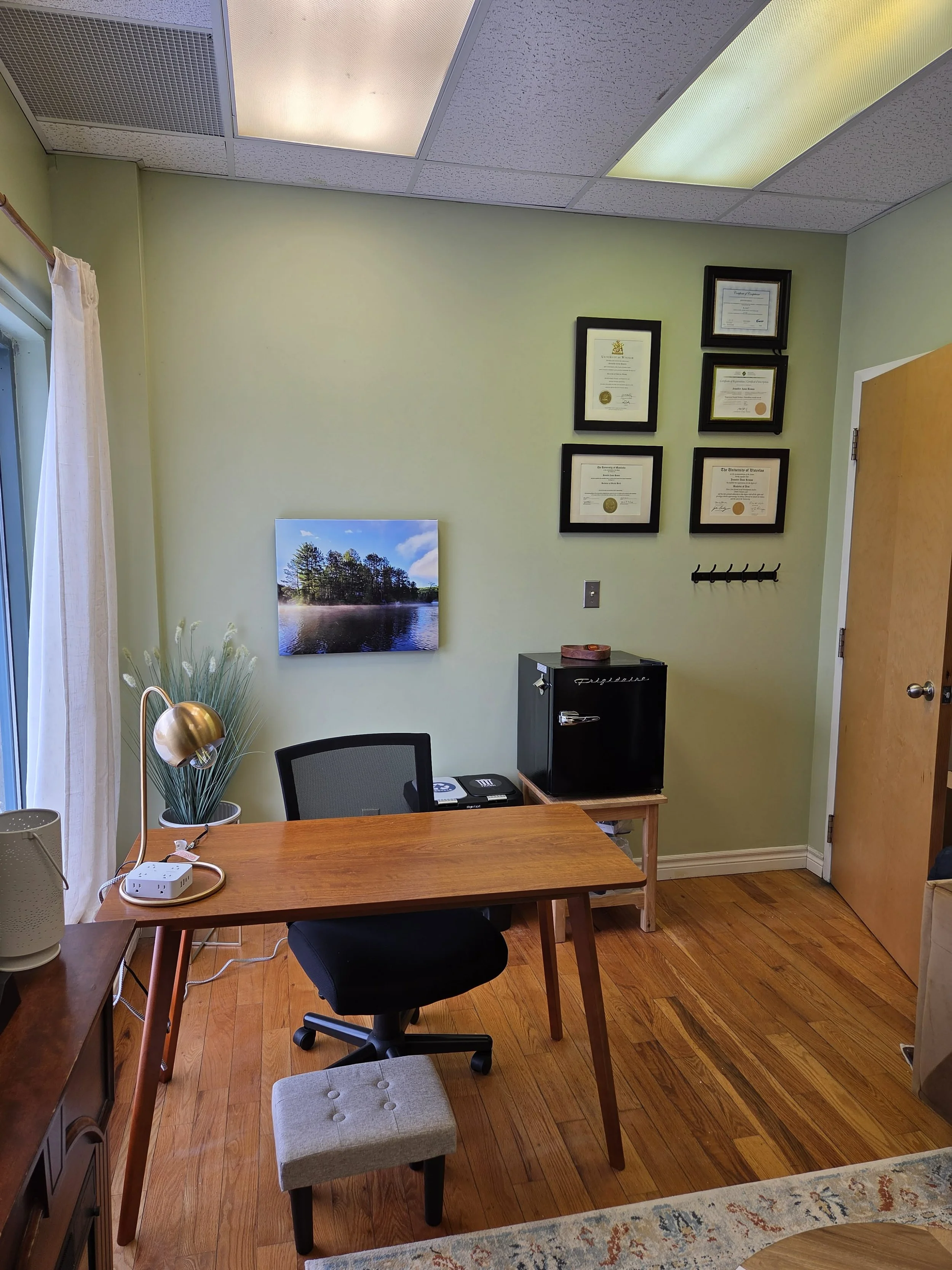 An office with a wooden desk and black office chair, taped pictures on the wall, a small black refrigerator, framed certificates, a window with curtains, and hardwood floor.