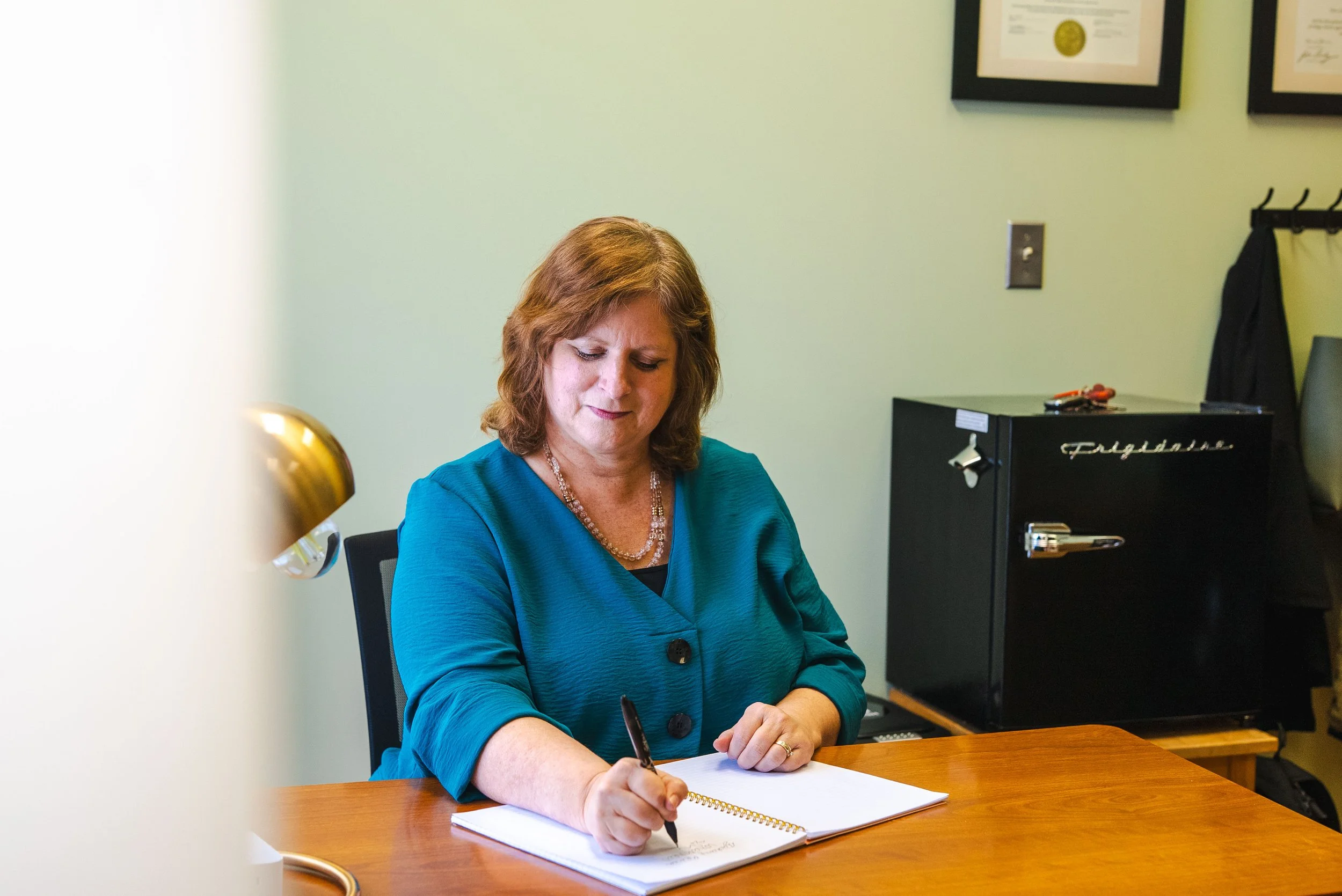Woman in blue top sitting at a desk, writing in a notebook, with framed certificates on the wall behind her.
