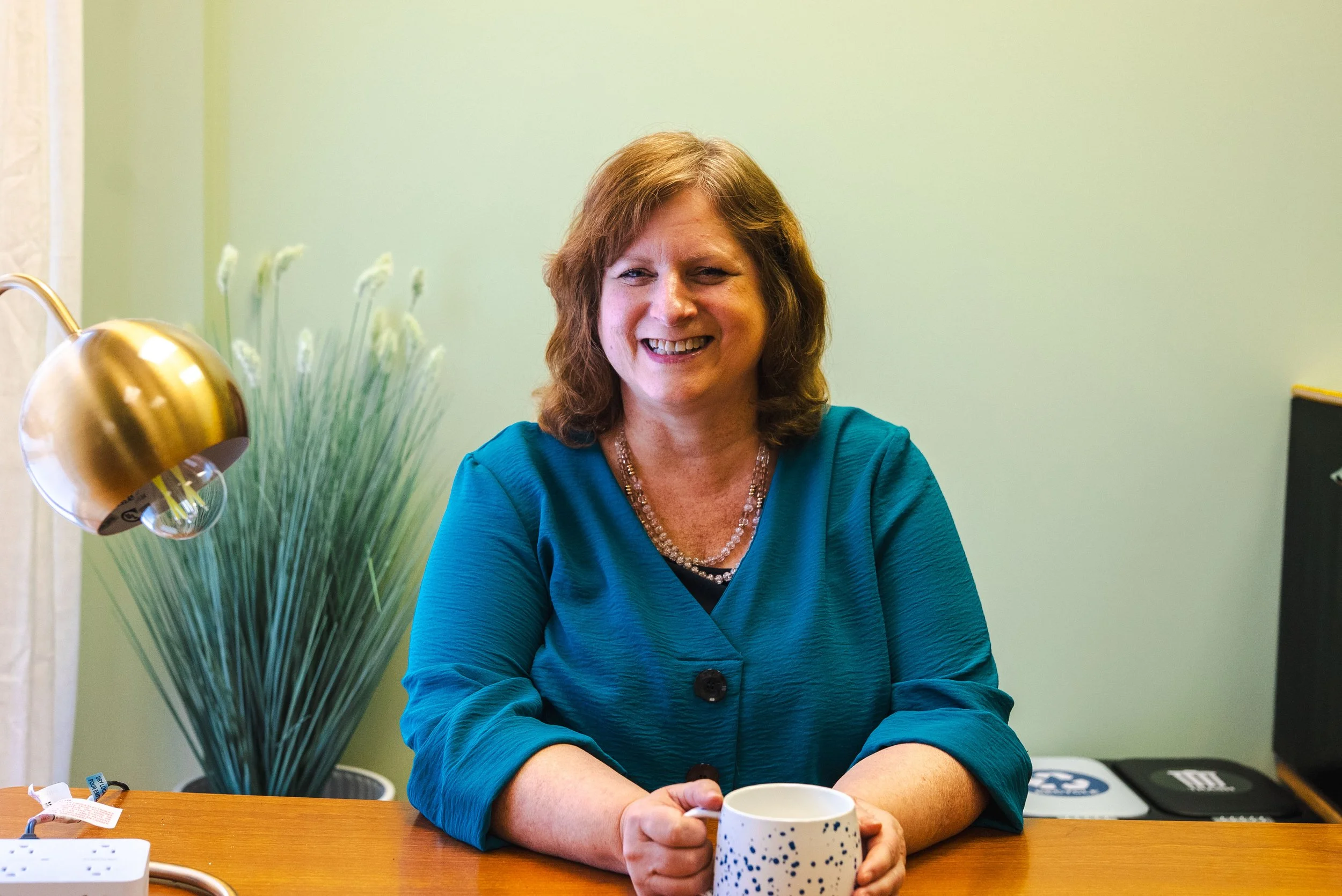 A woman with shoulder-length brown hair, smiling, sitting at a wooden desk. She is wearing a teal blouse and a beaded necklace, holding a speckled white and blue mug. In the background, there is a tall plant with white flowers and a gold desk lamp on the left, and part of an electronic device on the right.
