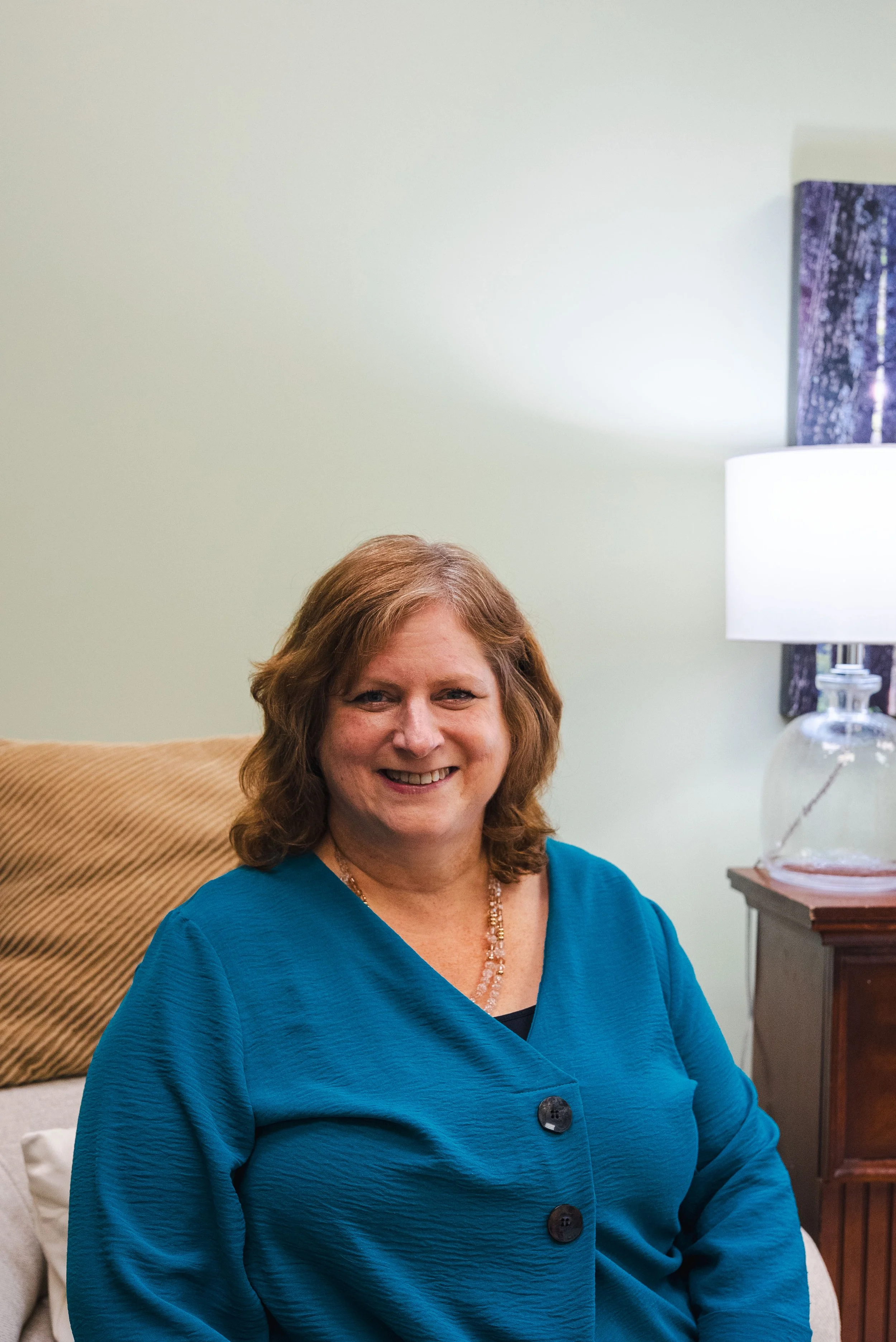 Smiling woman with reddish-brown hair, wearing a blue blouse and pearl necklace, sitting in a cozy room with a lamp and a painting in the background.