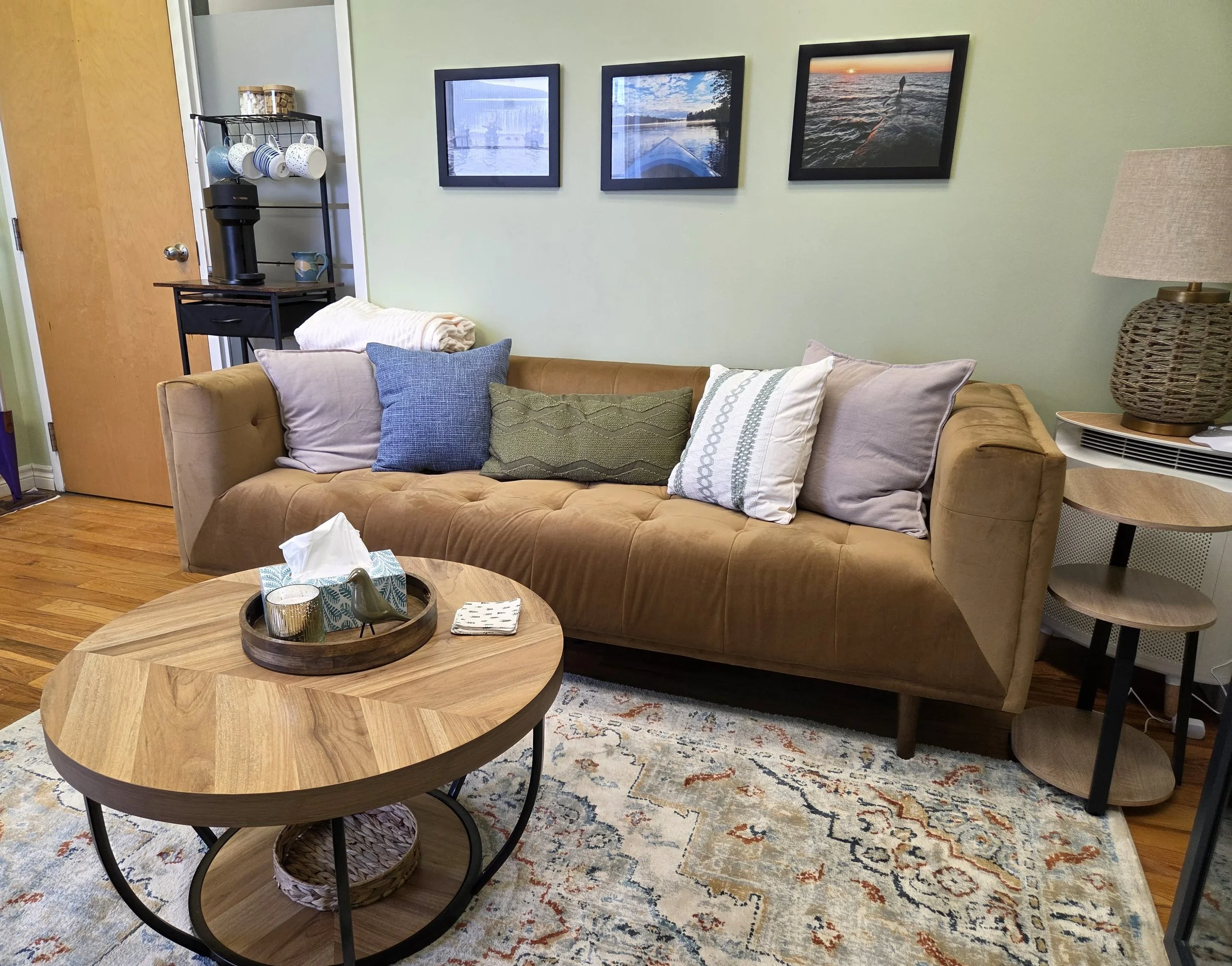 A living room with a tan couch adorned with six decorative pillows in various shades of blue, white, and green. A wooden round coffee table with a black metal base is in front of the couch, holding a tissue box, candles, and a small decorative bird. To the left of the couch, there is a black shelving unit with a coffee maker and cups. Above the couch, three framed landscape photographs are hung on a light green wall. There's a small side table with a woven lamp on the right side of the couch. The room features hardwood floors and a patterned area rug.