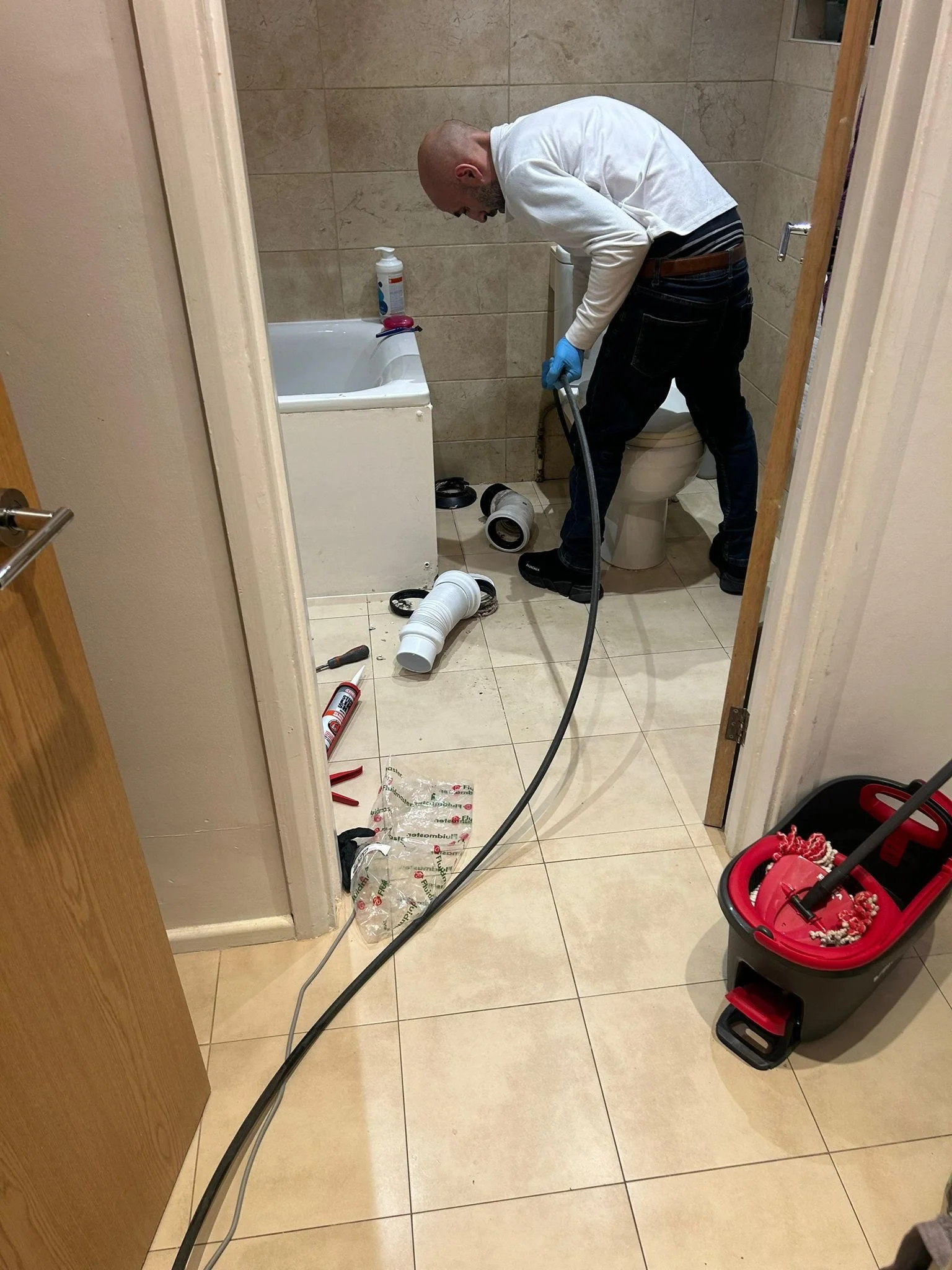 Under Pressure Plumbing engineer clearing a blocked bathroom waste pipe using a flexible drain-cleaning cable, with disconnected drainage pipes beside the bath and toilet.