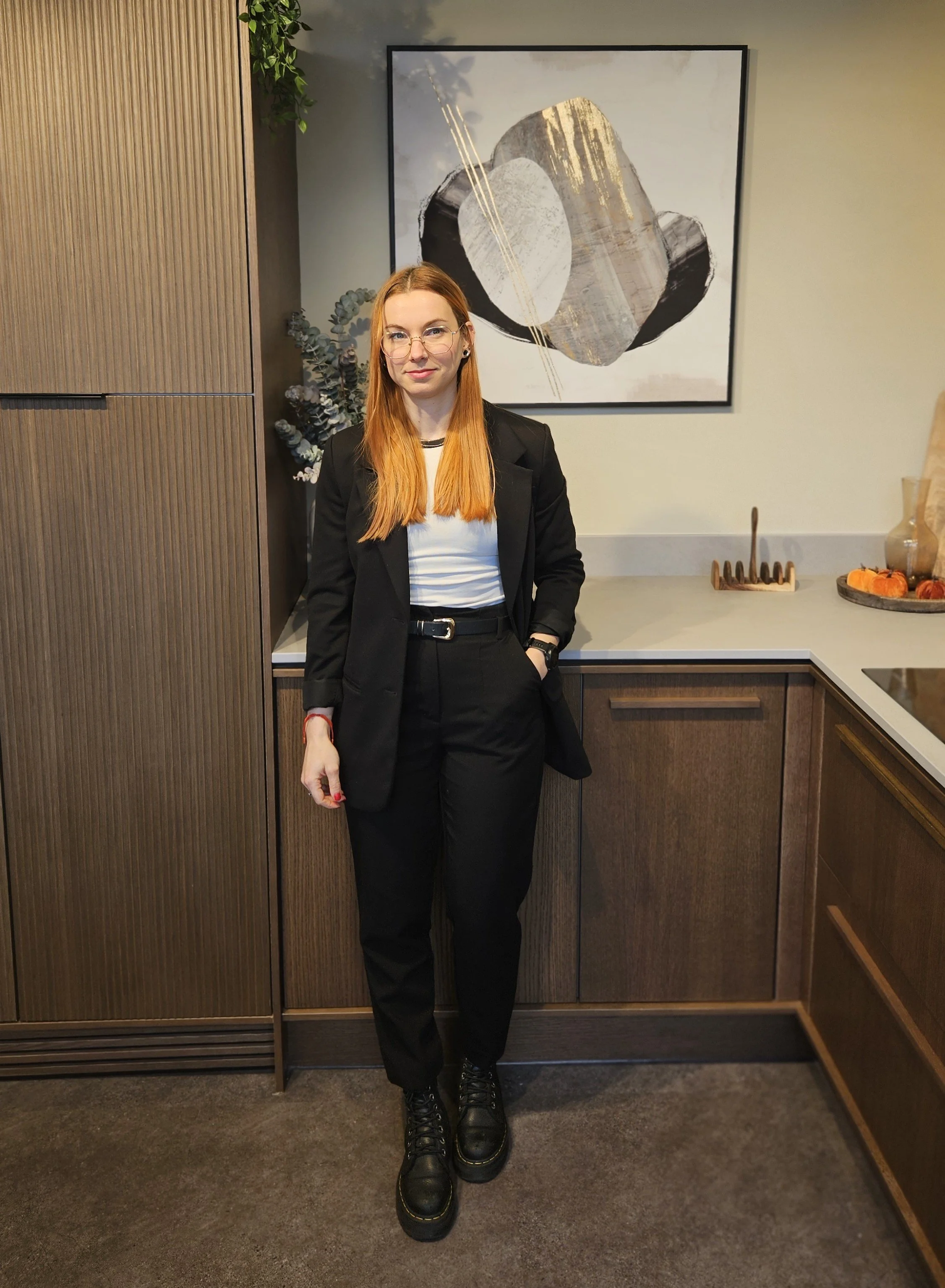 A woman with long red hair, wearing glasses, a black blazer, white shirt, black pants, and boots, standing in front of a modern abstract art piece in a Tasteful interior with wooden cabinets and decorative items.