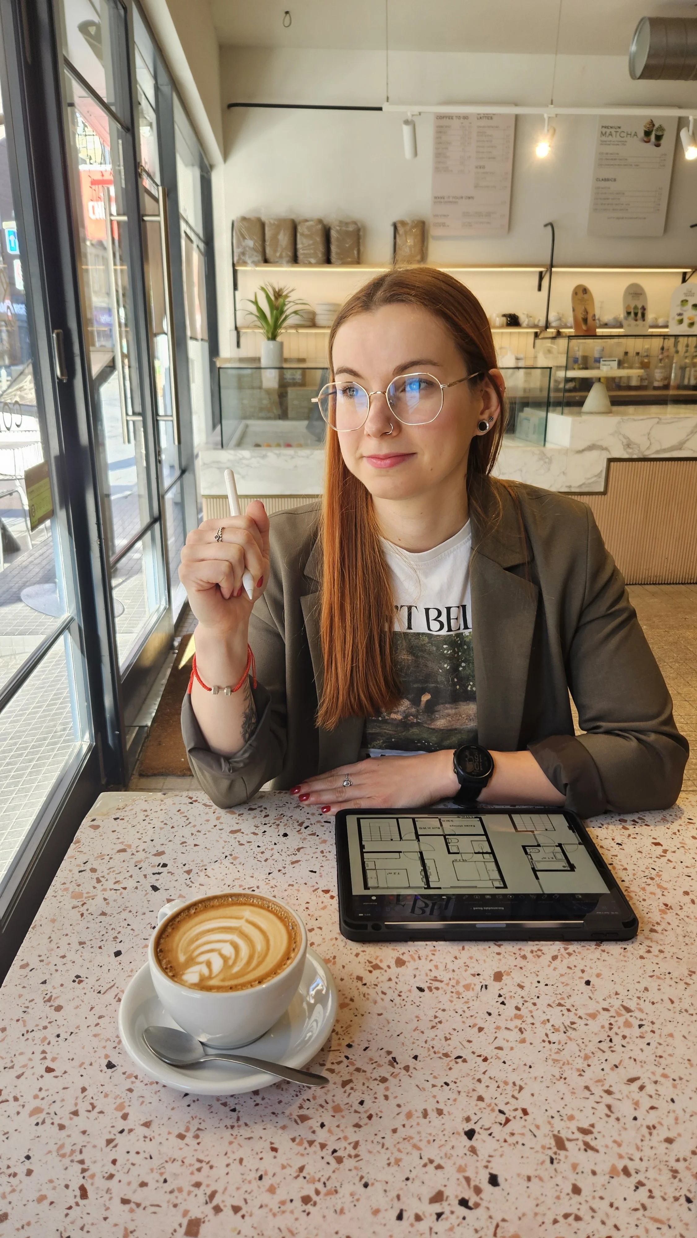 A young woman with long red hair, glasses, and piercings sitting at a cafe table. She is holding a pen and looking to the side. On the table in front of her is a cup of latte with latte art and a tablet displaying a floor plan.