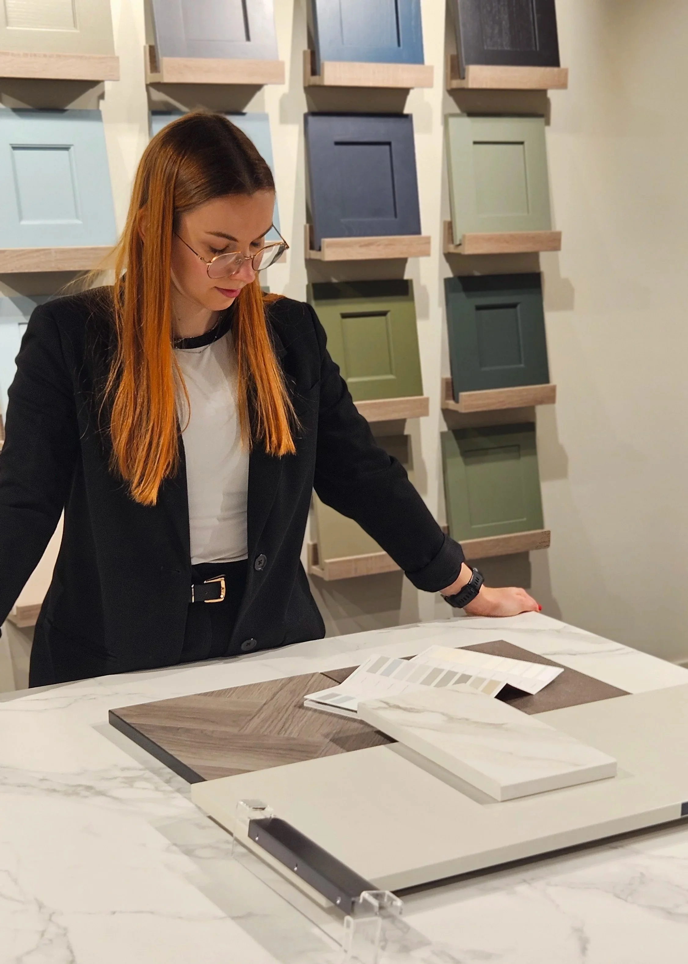 A woman with long red hair, wearing glasses, a black blazer, and a white shirt, looking at color and material samples displayed on a marble table in a showroom or interior design store.