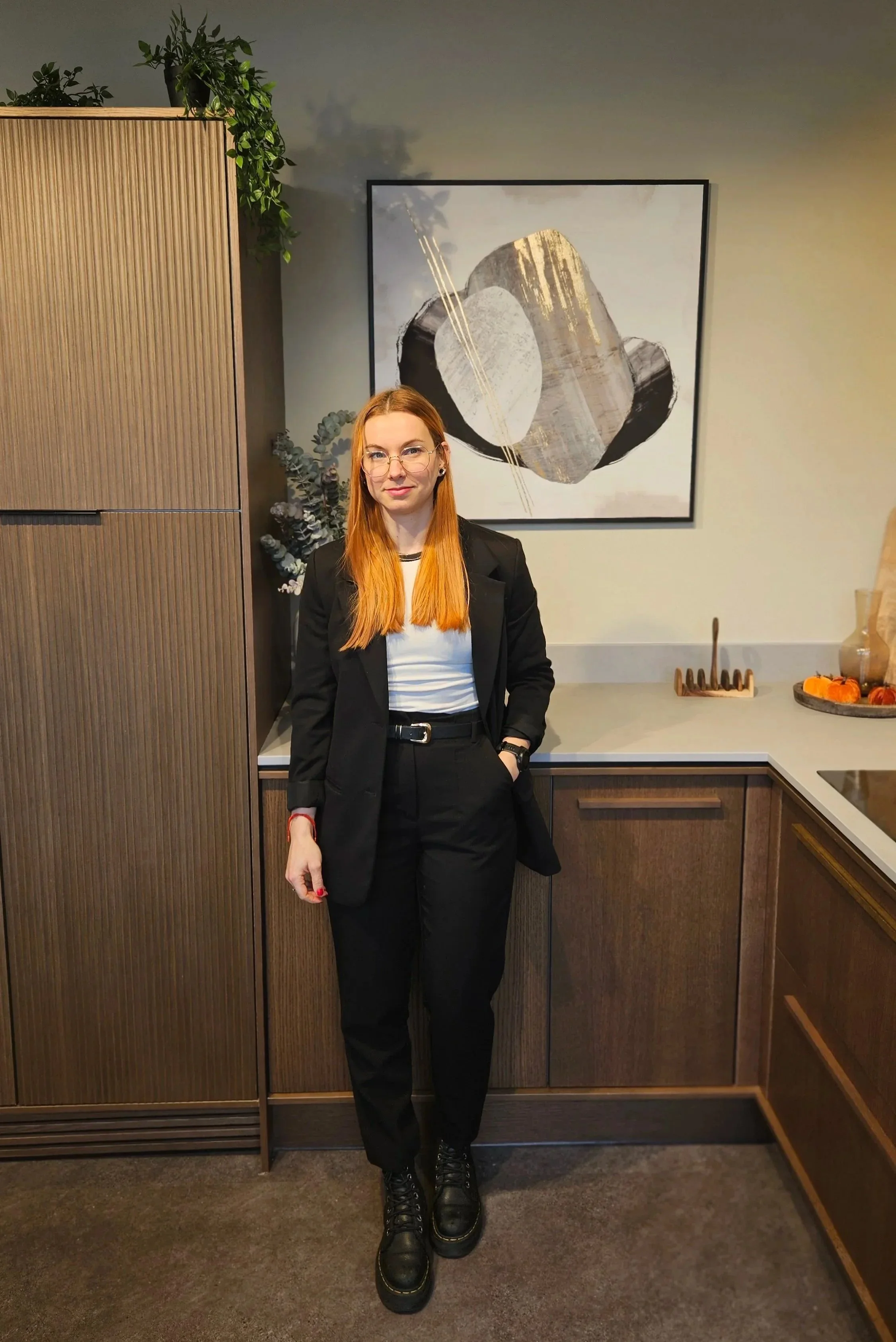 A woman with long red hair, glasses, and a black suit standing in a modern kitchen with wood cabinets, abstract wall art, and decorative plants.