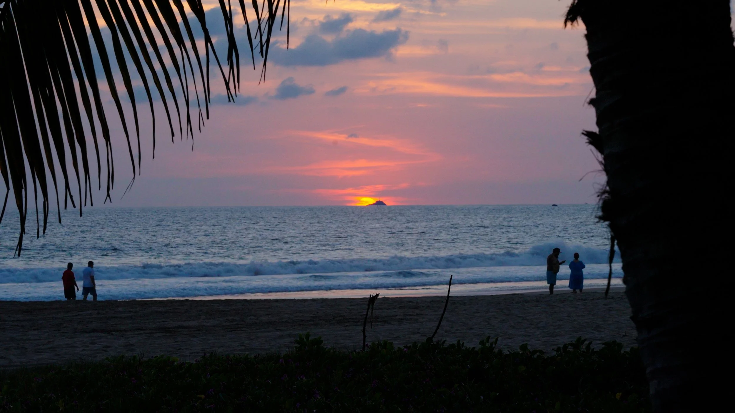 Sunset over the ocean viewed from a beach, with silhouettes of two couples walking and talking on the sand, framed by palm trees.