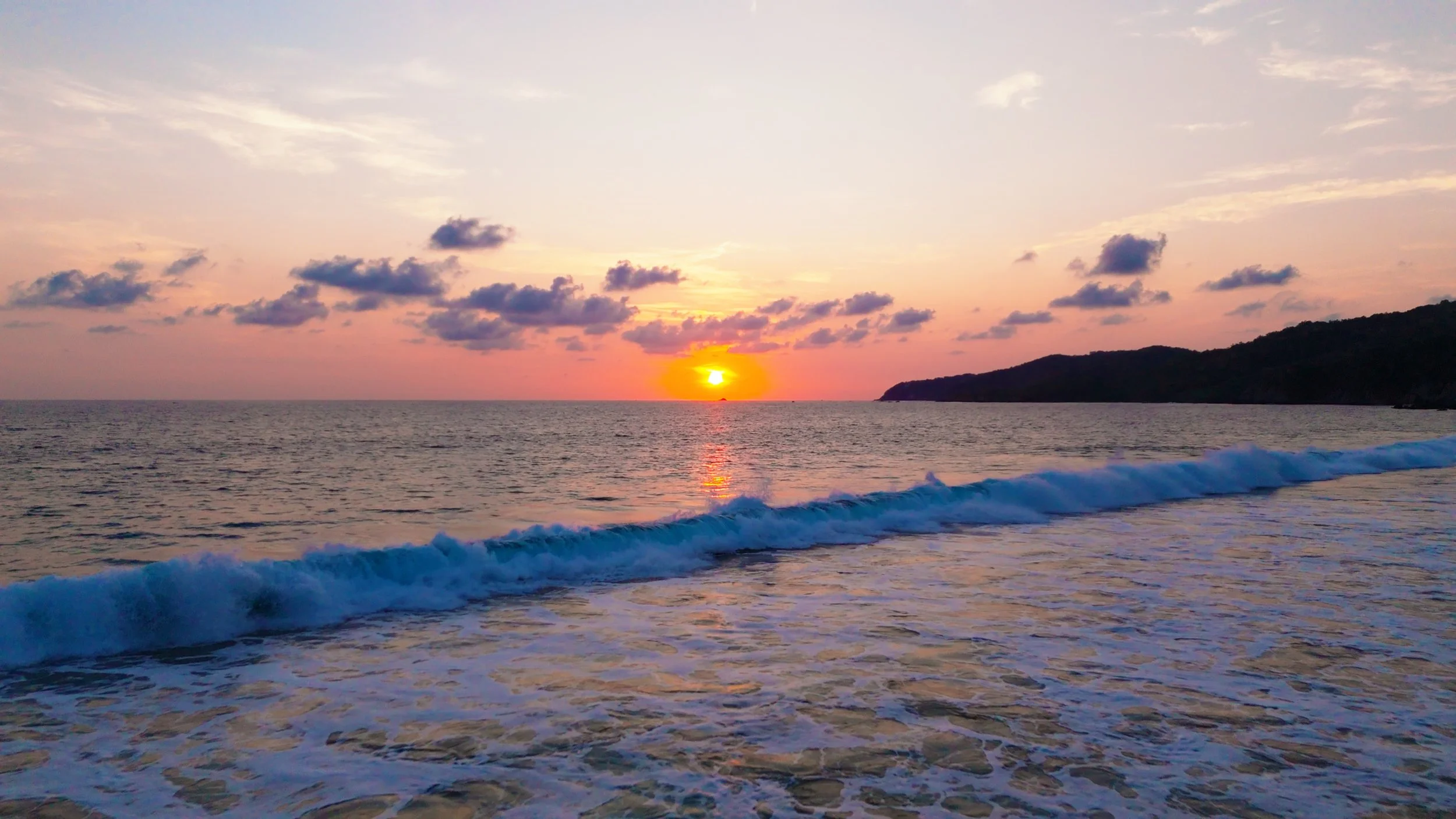Sunset over the ocean with waves crashing on the shore, clouds in the sky, and a silhouette of a landmass in the distance.