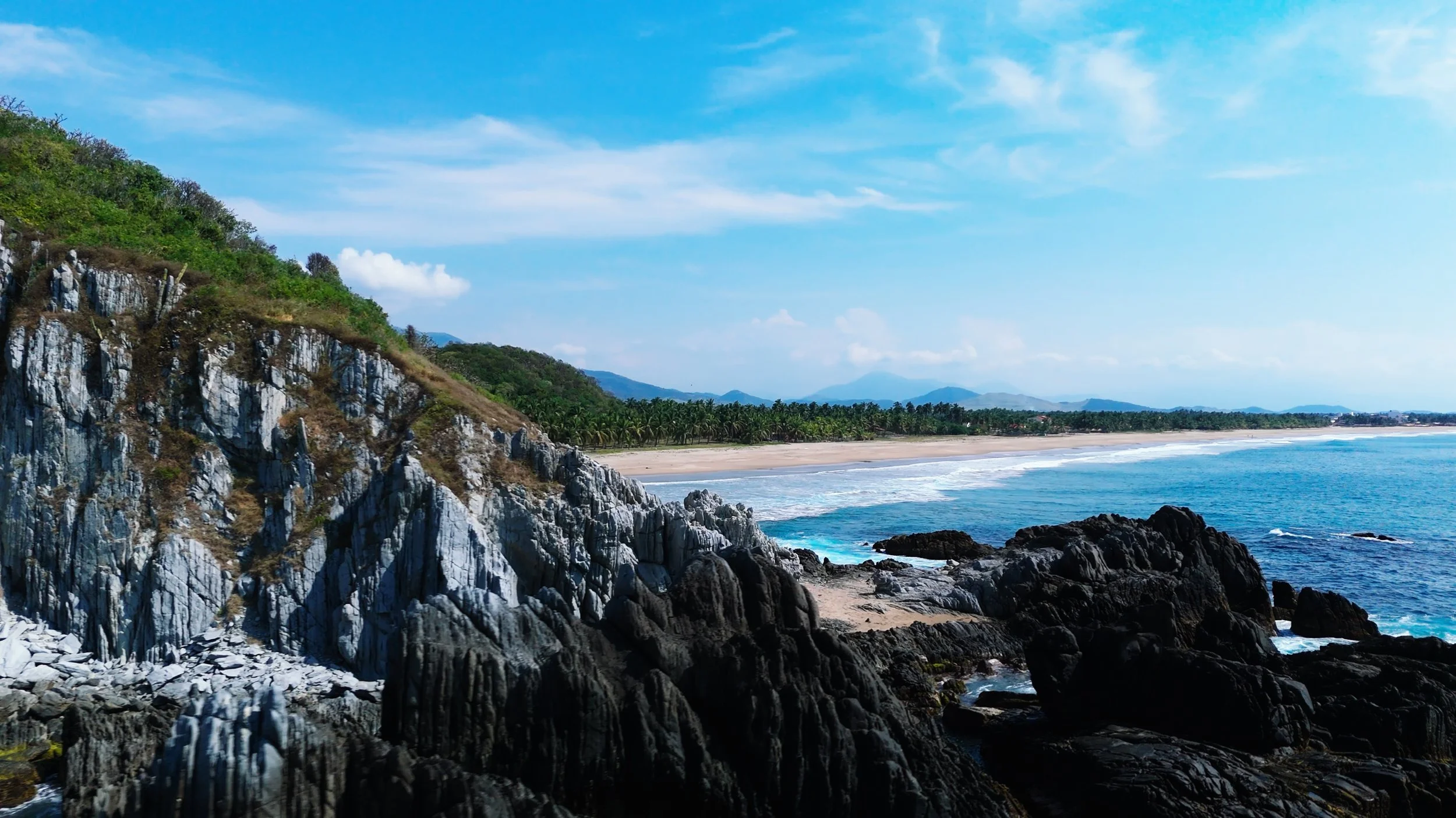 A scenic coastal view with rocky cliffs on the left, a sandy beach with small waves in the middle, and a line of palm trees with mountains in the background under a bright blue sky with scattered clouds.