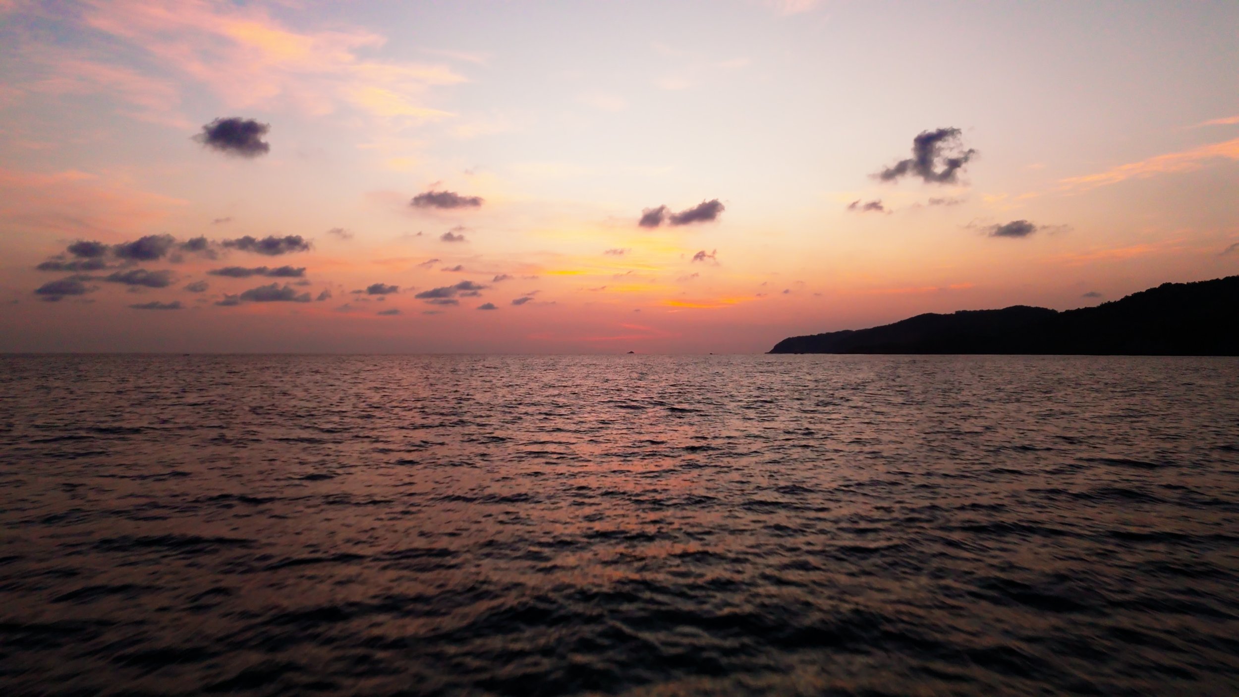 A sunset over the ocean with a partly cloudy sky and a silhouette of a small landmass on the horizon.