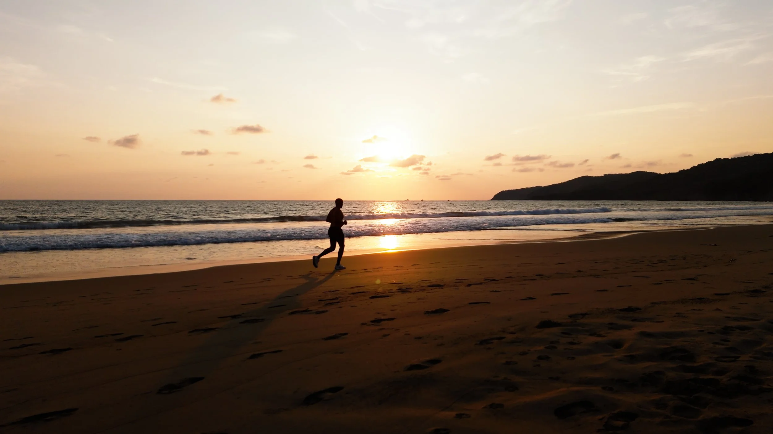 Person jogging on a beach during sunset with the ocean waves and a distant landmass in the background.