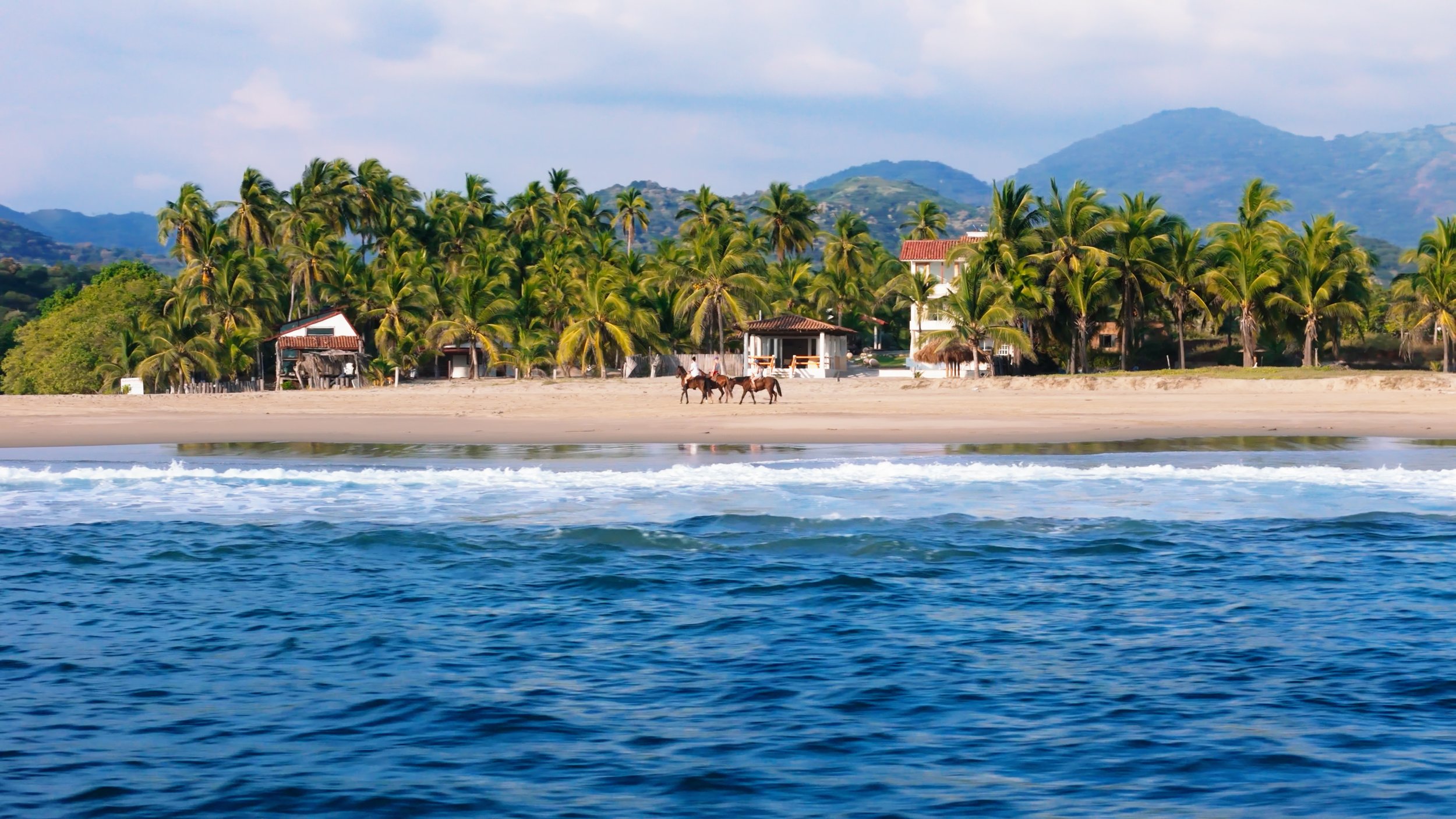 Beach scene with blue water in foreground, sandy shore with palm trees and small buildings or huts, and mountains in the background, under partly cloudy sky.