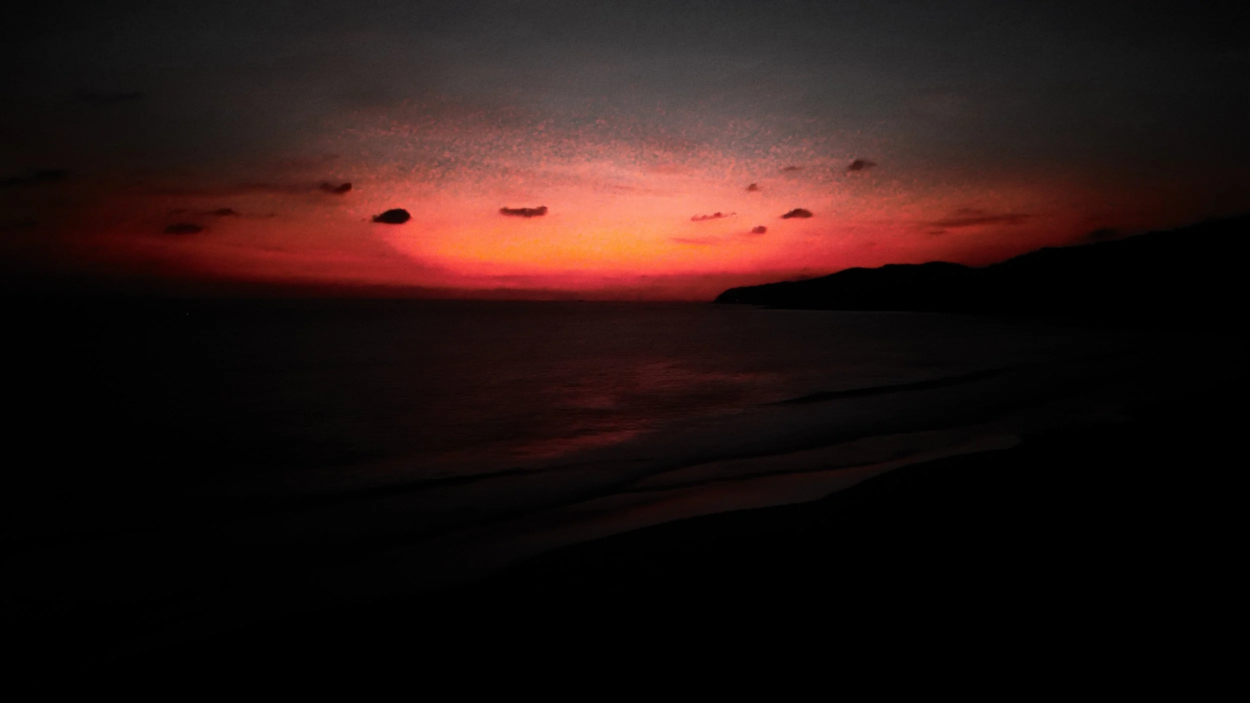 Sunset over the ocean with pink and purple hues in the sky, silhouette of hills and dark shoreline.