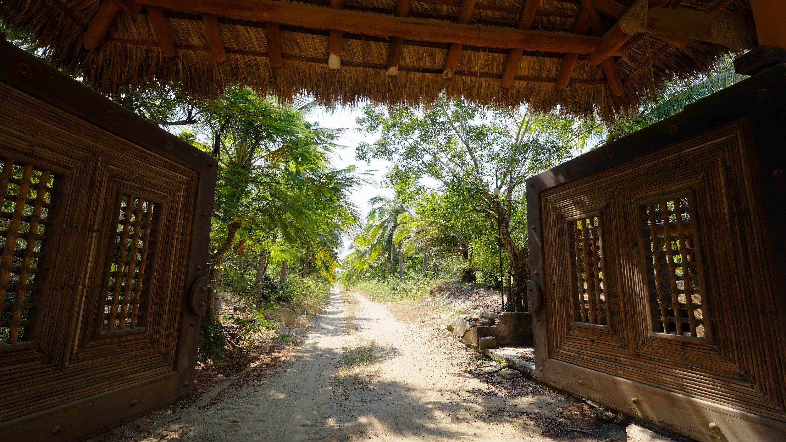 View of a sandy pathway through tropical trees, seen from behind wooden gate doors, with a thatched roof overhang in the foreground.