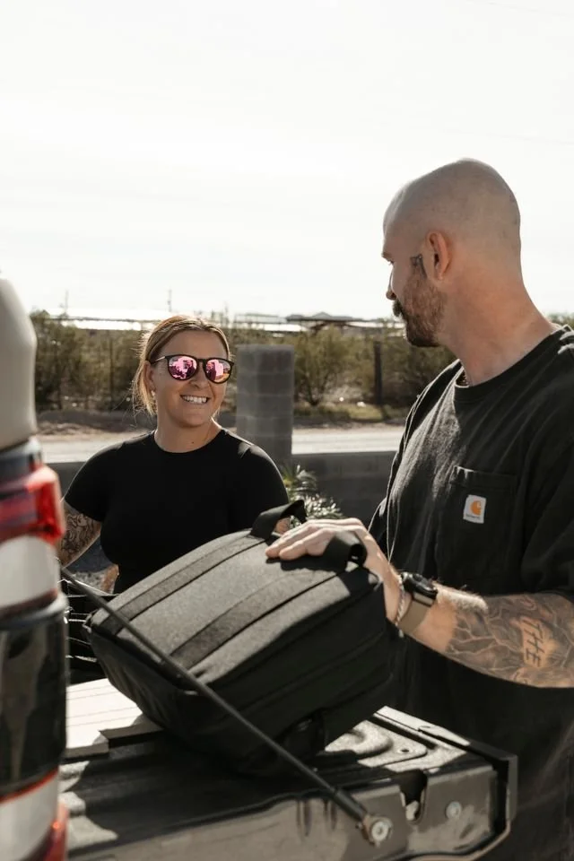 A woman with sunglasses and a black shirt smiling and talking to a man with a shaved head, beard, black Carhartt shirt, and tattoos, standing near the back of a vehicle with a gray backpack on the tailgate.