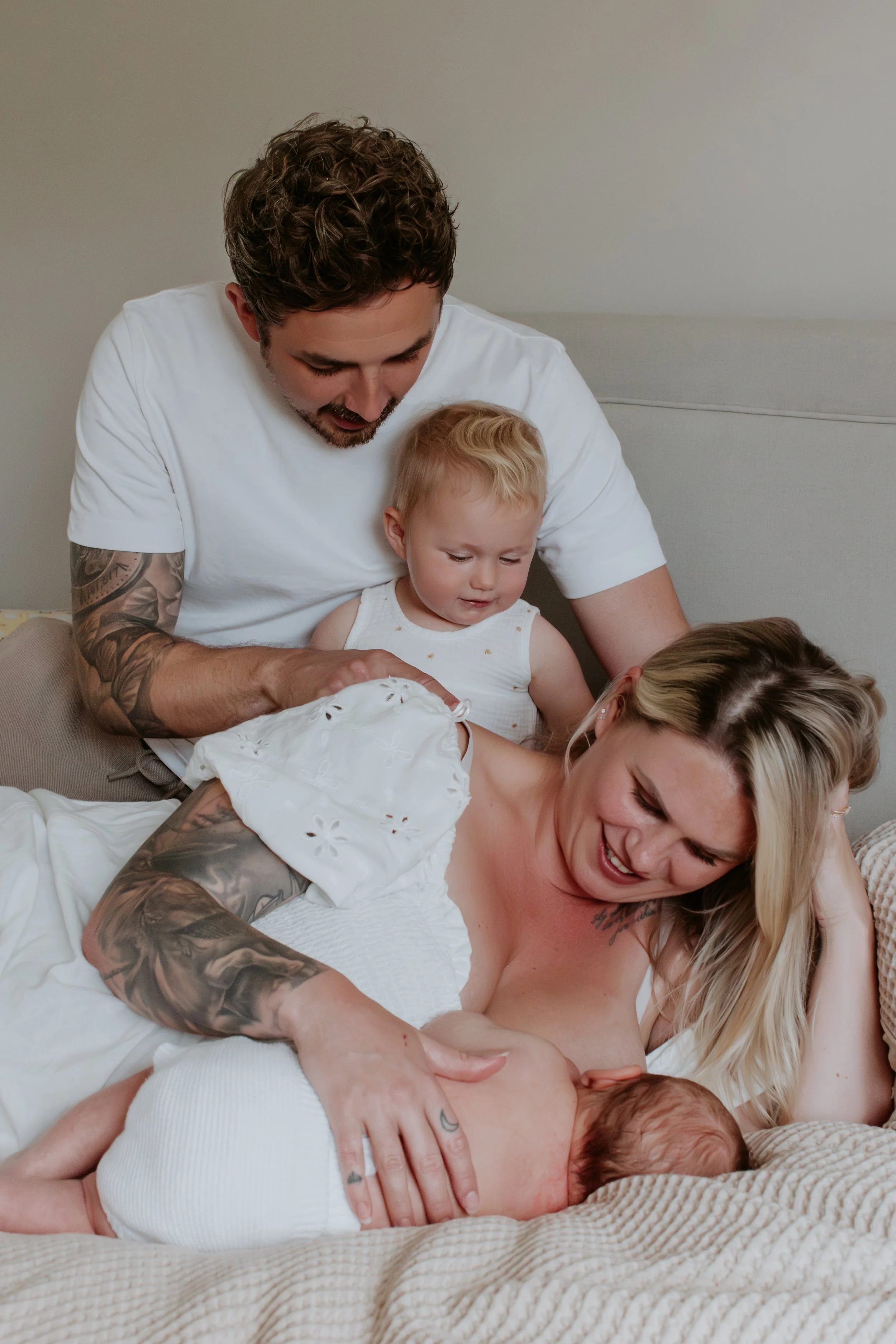A family of four with tattoos, including a woman breastfeeding a newborn, a man, and two young children, gathered on a bed in a cozy bedroom.