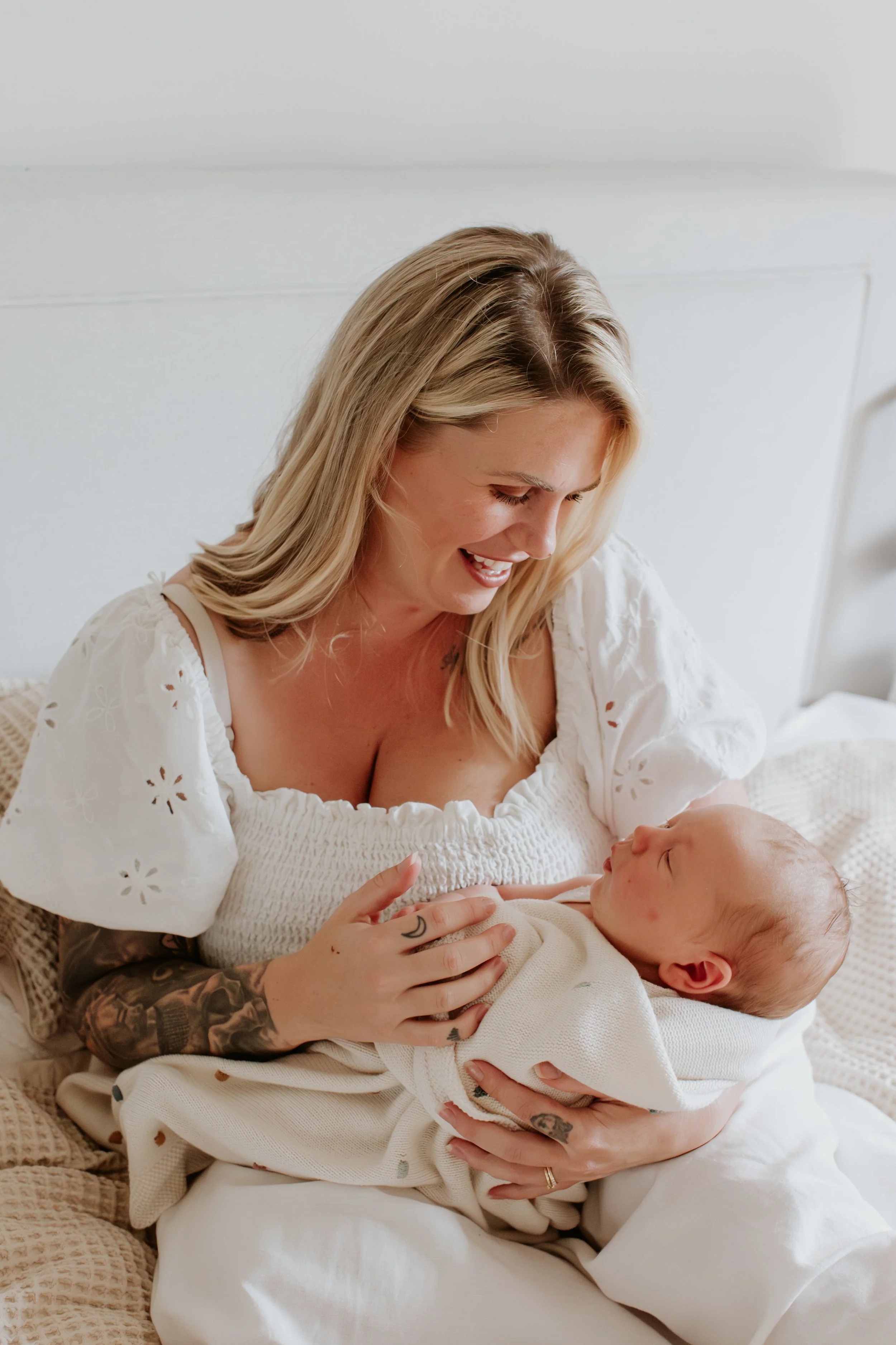 A woman with blonde hair holding a baby while sitting on a bed, both smiling and looking at each other in a bright room.
