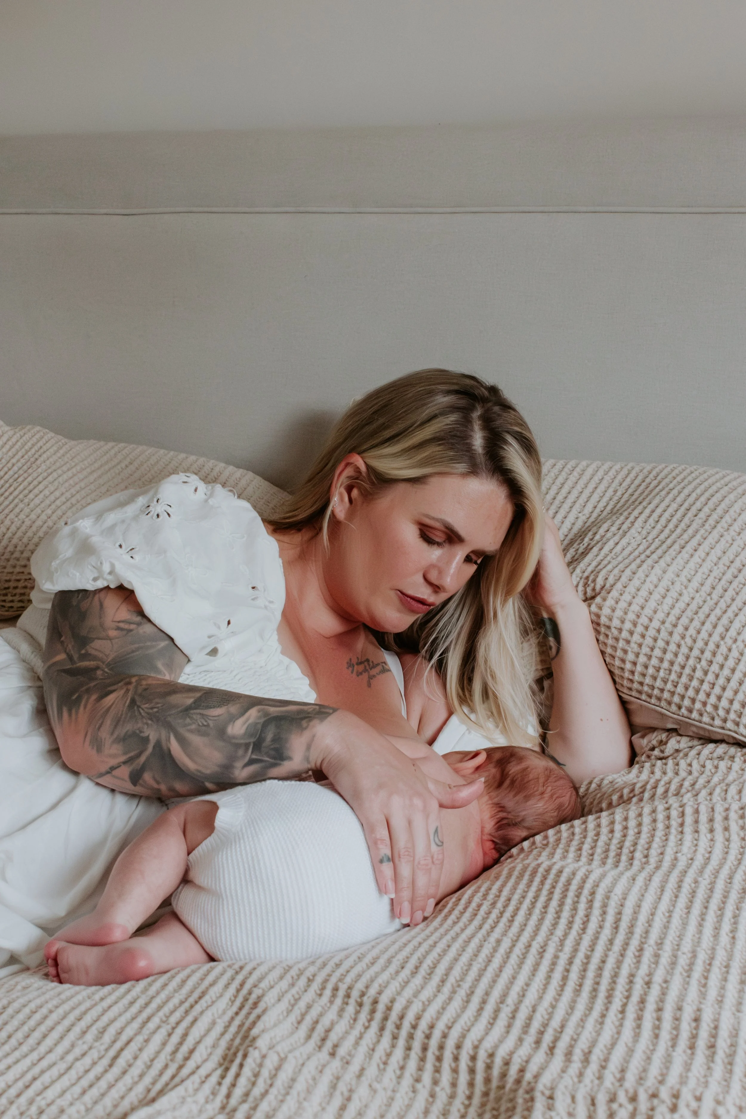A woman with blonde hair and tattoos on her arms is lying on a beige bed, breastfeeding a newborn baby wrapped in a white blanket.