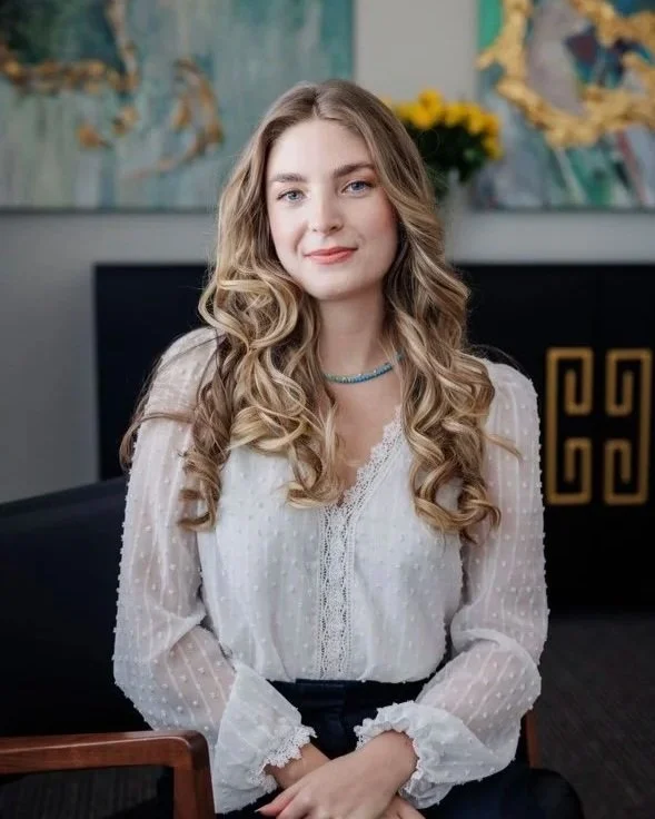 A young woman with long, curly blonde hair, wearing a white blouse with lace and polka dots, sitting in a modern room with artwork and flowers in the background.