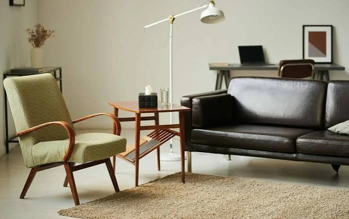 Living room with a green armchair, a wooden side table with tissue box and glasses, black leather sofa, floor lamp, and a desk with a laptop and framed picture in the background. NC SC VA Madison Long trauma EMDR pregnancy postpartum eating disorders