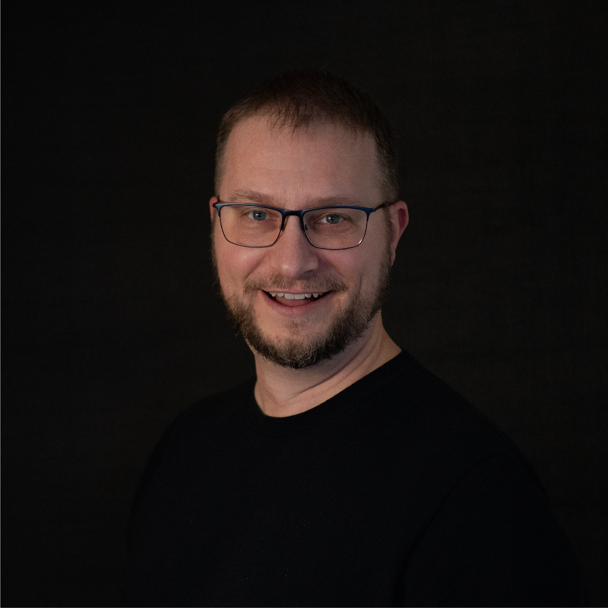 A smiling man with glasses, a beard, and short brown hair, wearing a black shirt against a dark background.