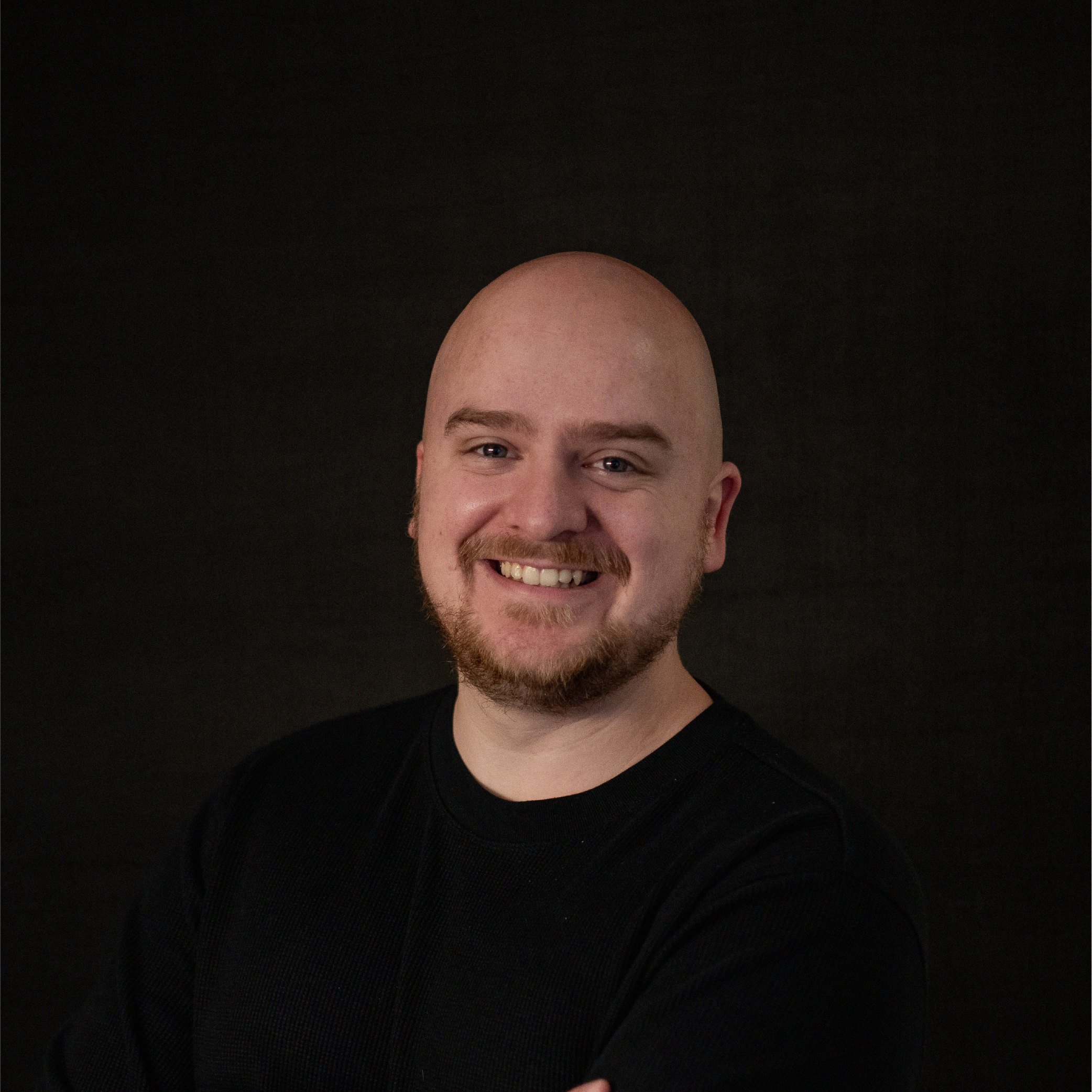 A smiling bald man with a beard and mustache wearing a black shirt, posing against a dark background.