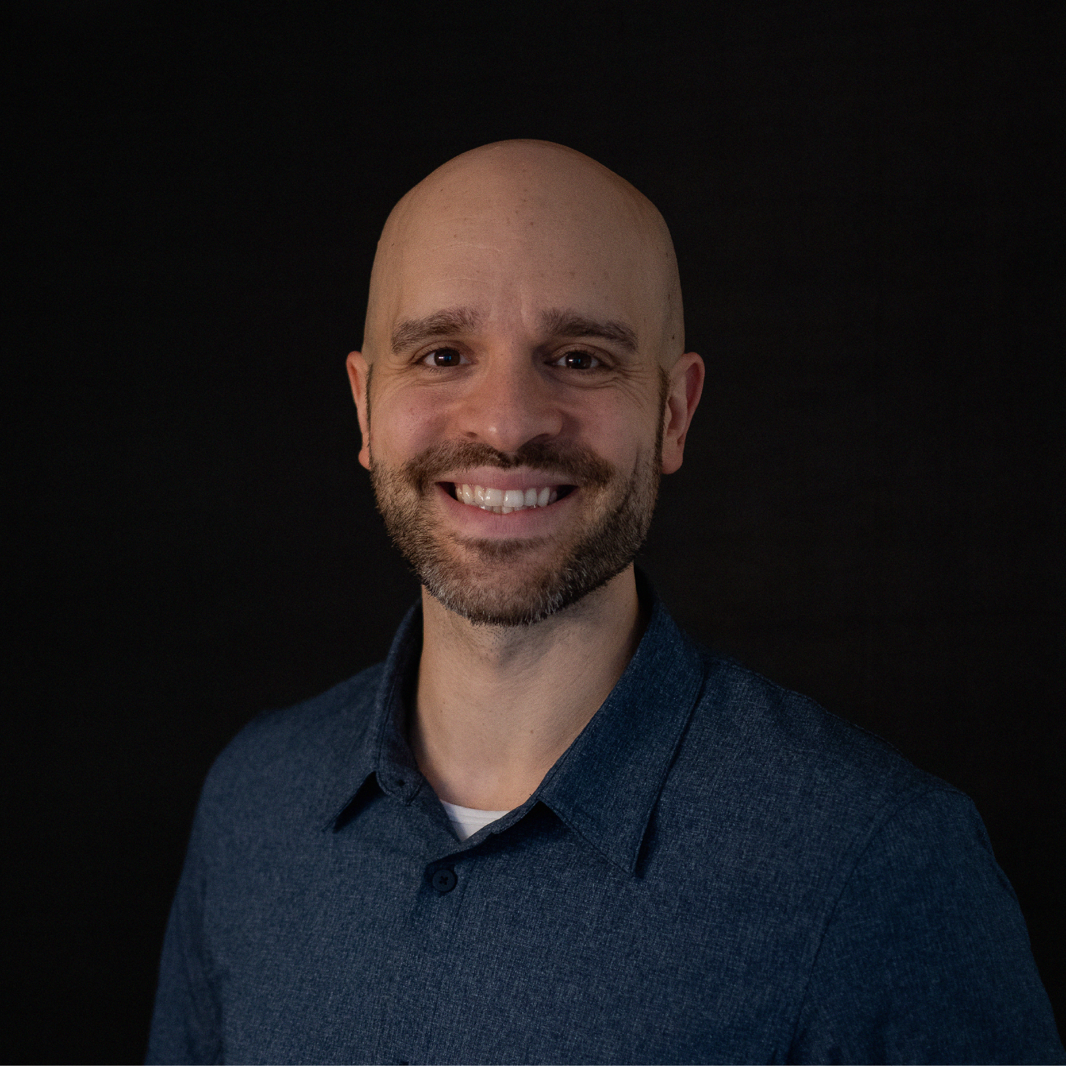 Portrait of a smiling bald man with a beard wearing a dark blue collared shirt against a black background.