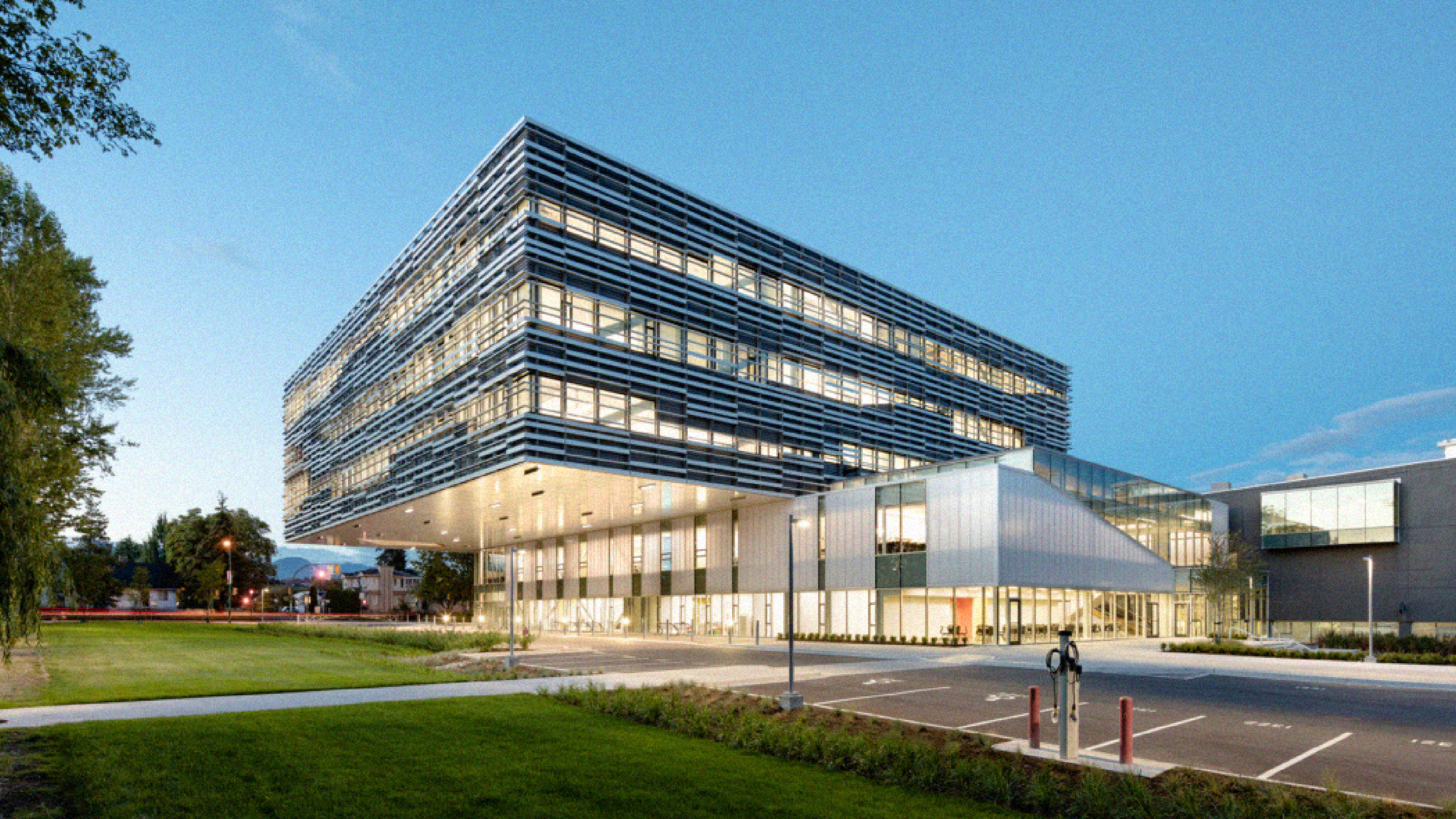 Modern multi-story building with glass and metal exterior, illuminated interior, parking lot, and bike station in foreground, surrounded by green lawn and trees, during twilight.