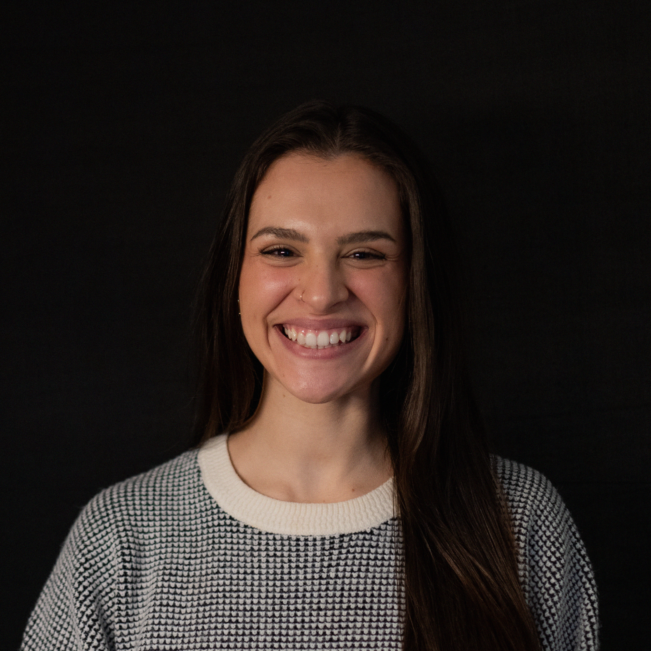 Portrait of a young woman with long brown hair, smiling and showing her teeth, wearing a patterned sweater, against a dark background.