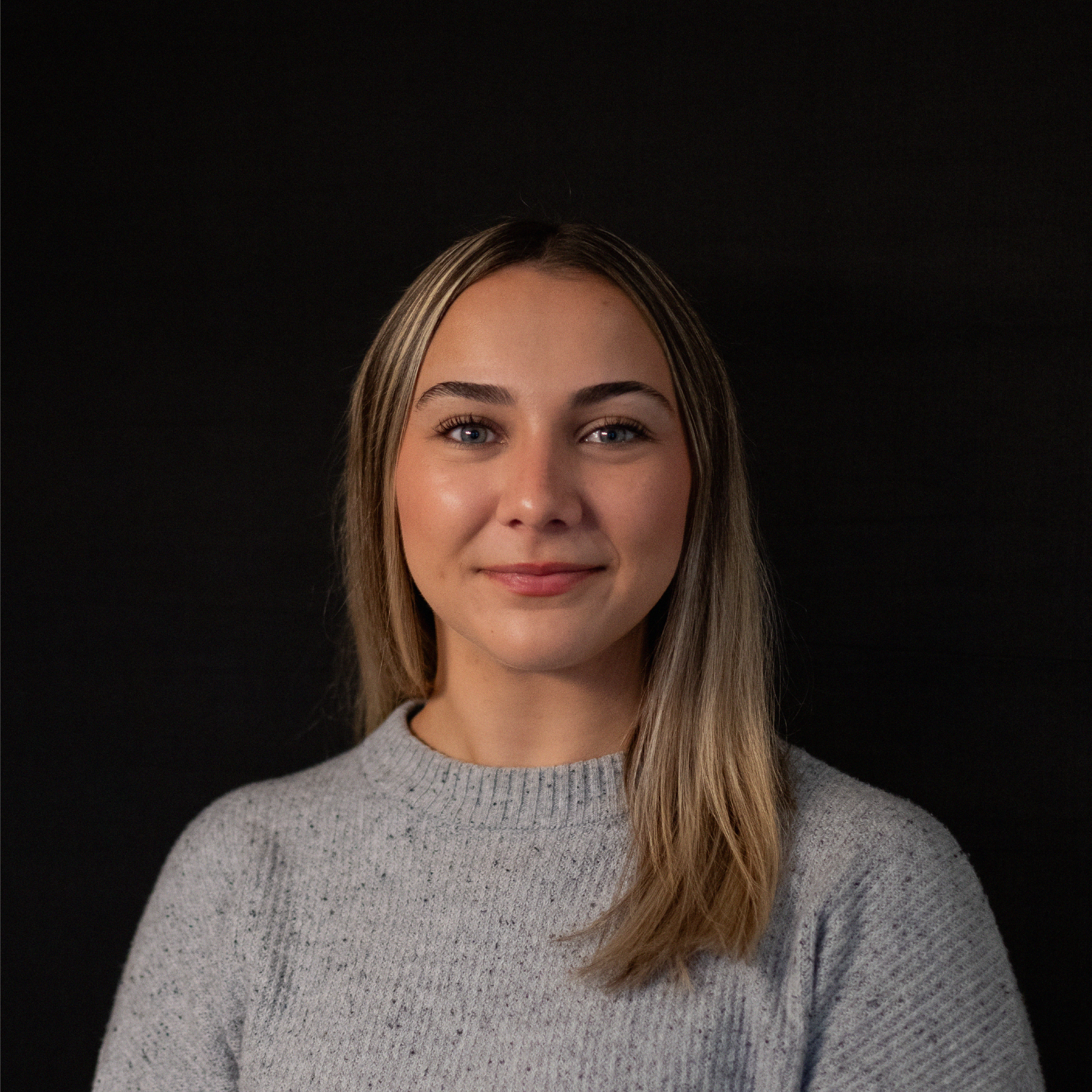 Portrait of a young woman with long blonde hair, blue eyes, and a slight smile, wearing a light gray sweater, against a solid black background.