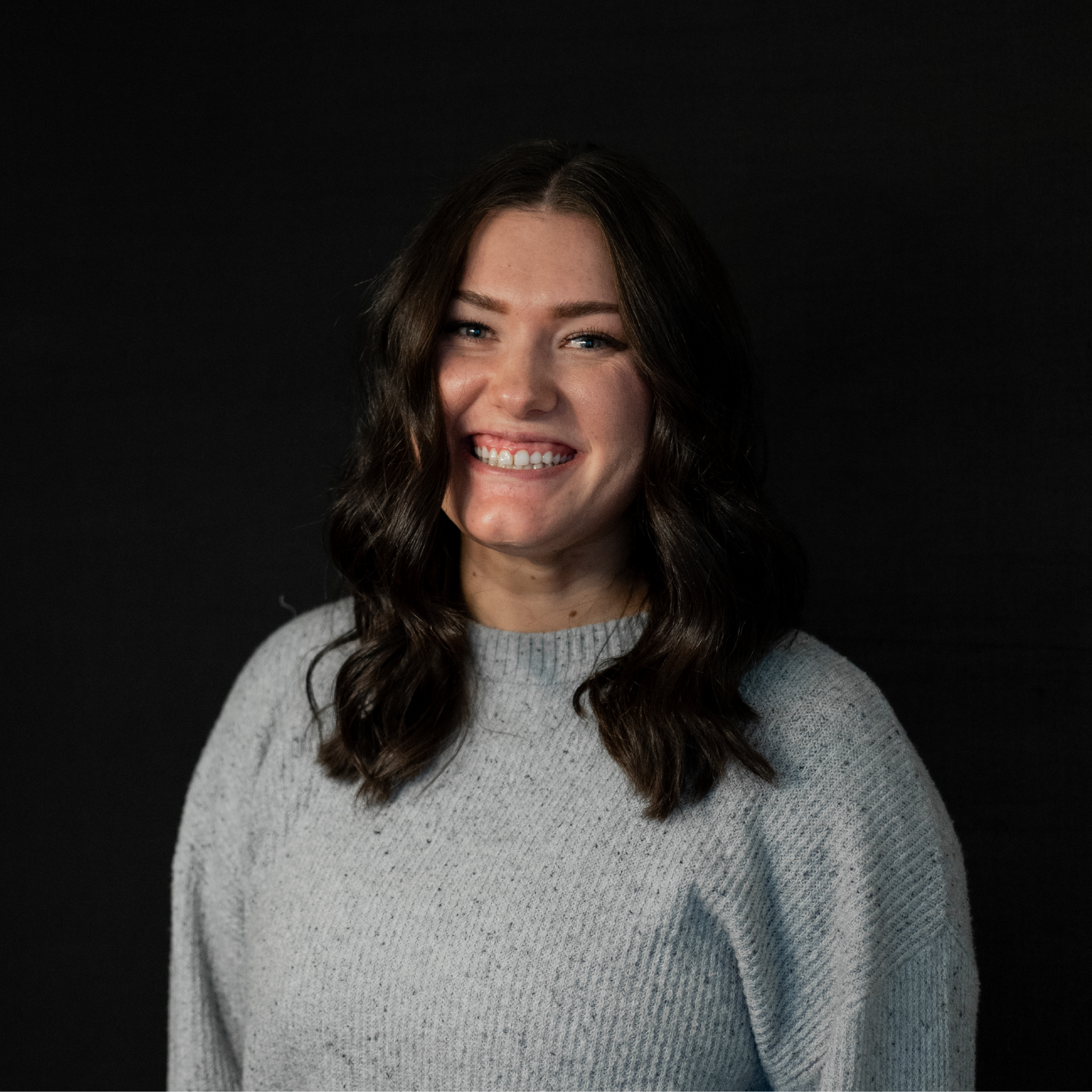Portrait of a smiling woman with dark wavy hair wearing a light gray sweater against a black background.
