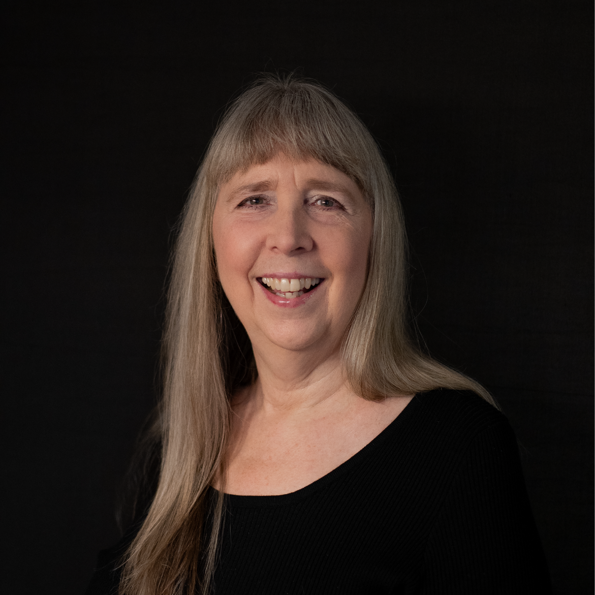 A smiling middle-aged woman with long gray hair and light skin, wearing a black top, against a dark background.