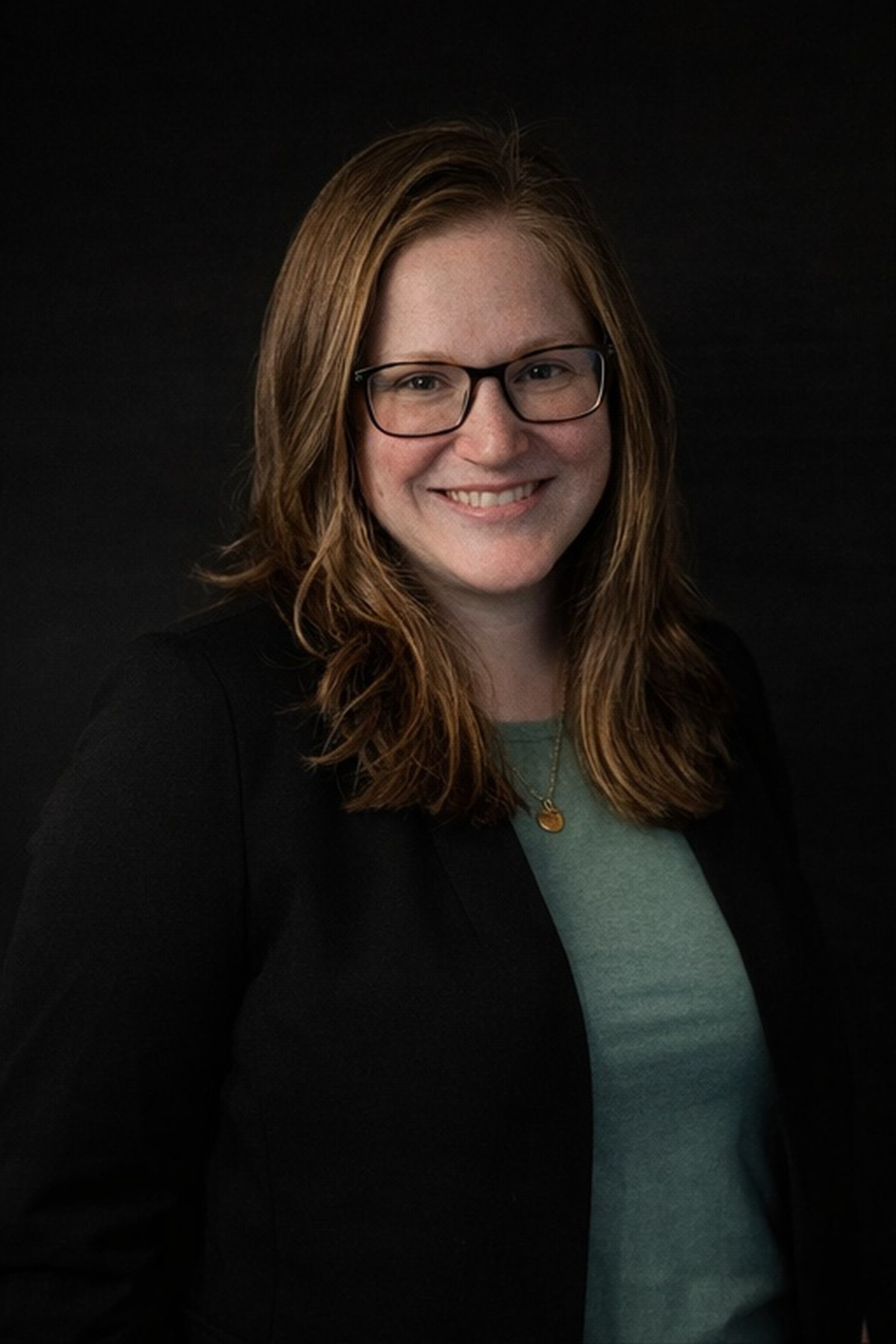 Portrait of a woman with shoulder-length brown hair, glasses, wearing a black blazer and a green top, smiling against a dark background.