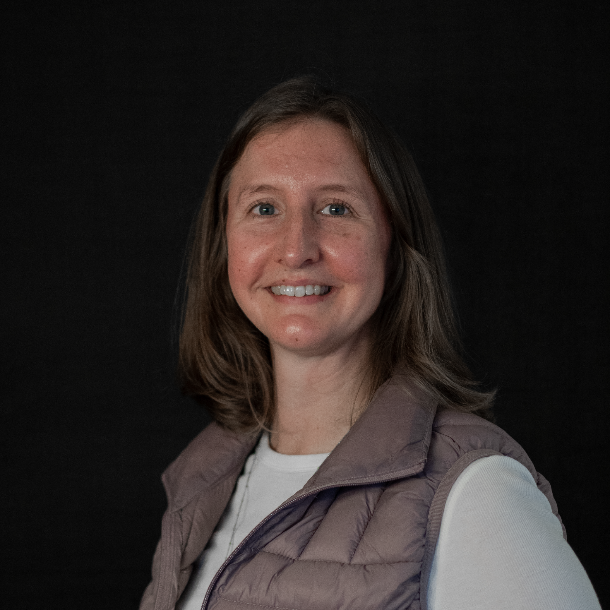 Portrait of a smiling woman with shoulder length brown hair, wearing a white shirt and a light purple vest against a black background.