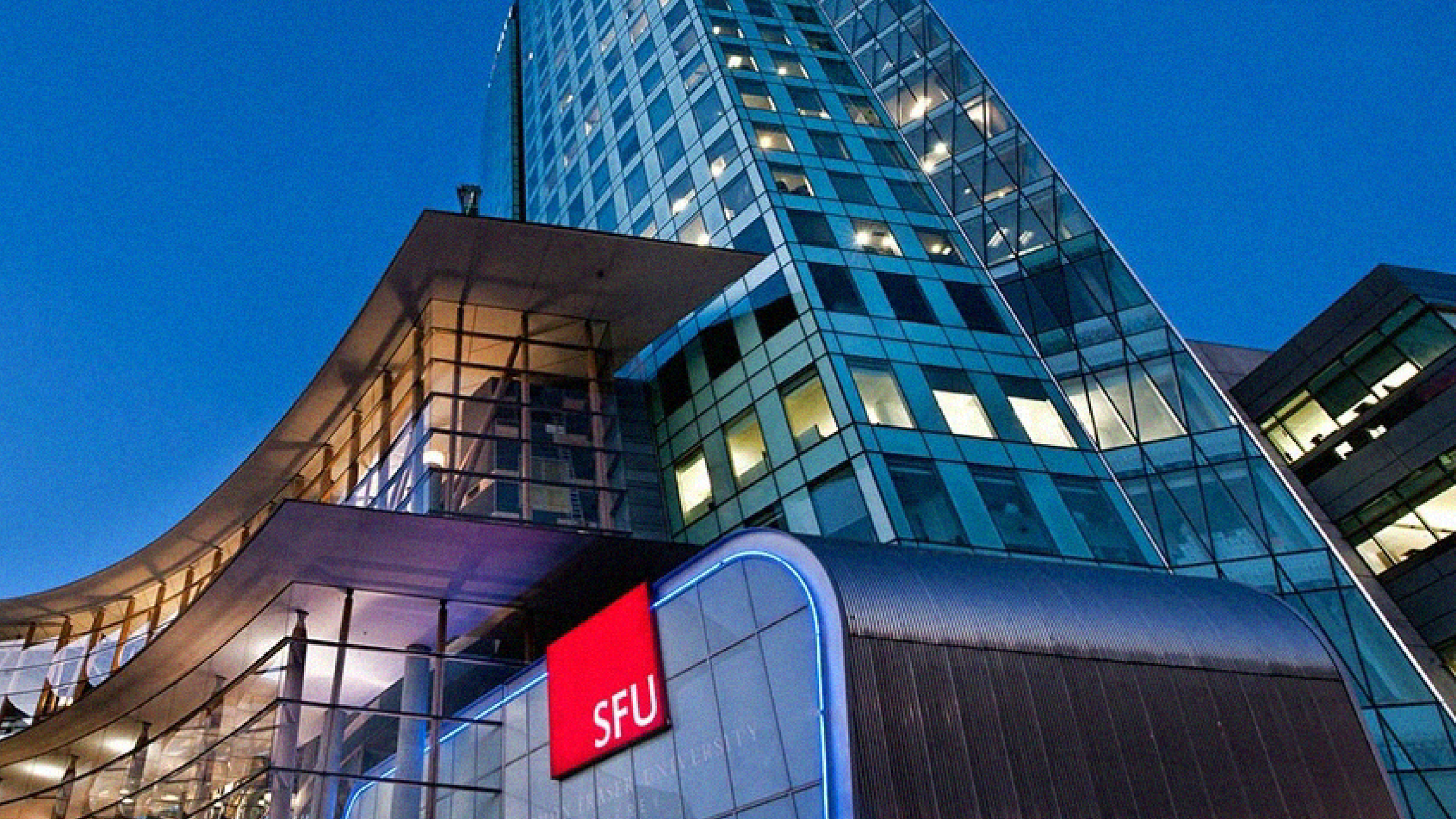 Nighttime view of a modern glass office building with illuminated windows and a sign reading SFU on the lower part.