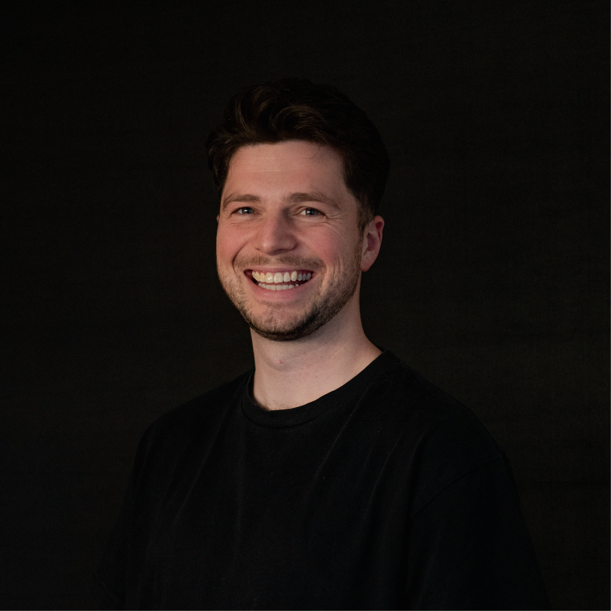 A young man with dark hair and a beard smiling against a black background.