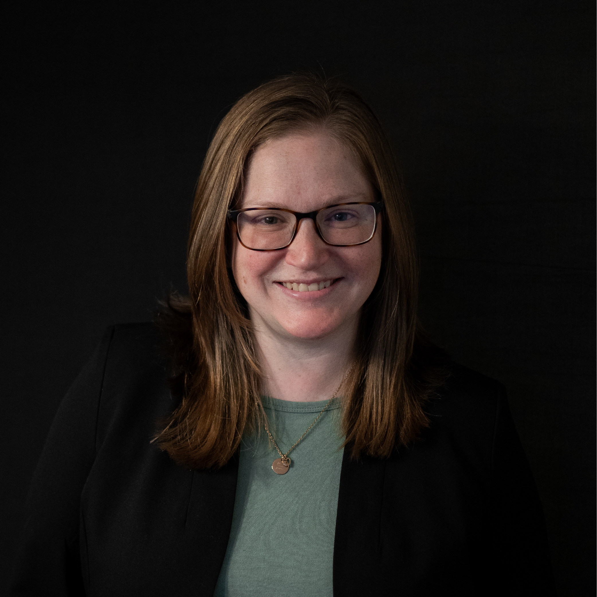Portrait of a woman with shoulder-length brown hair, glasses, wearing a black blazer and a green top, smiling against a dark background.