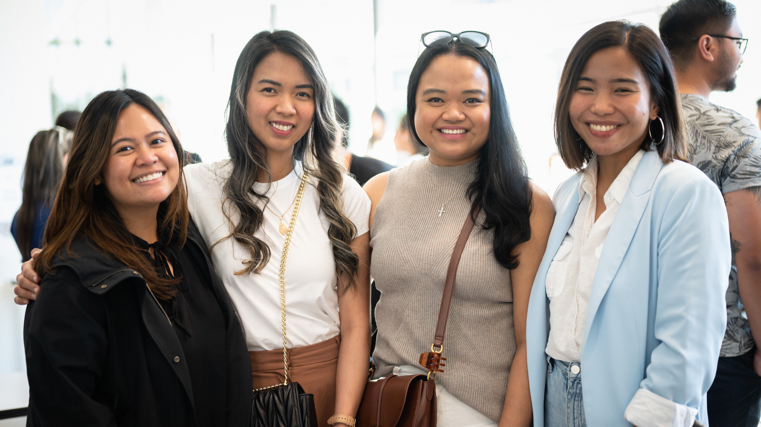 Four women smiling and standing close together in an indoor setting, with other people in the background.