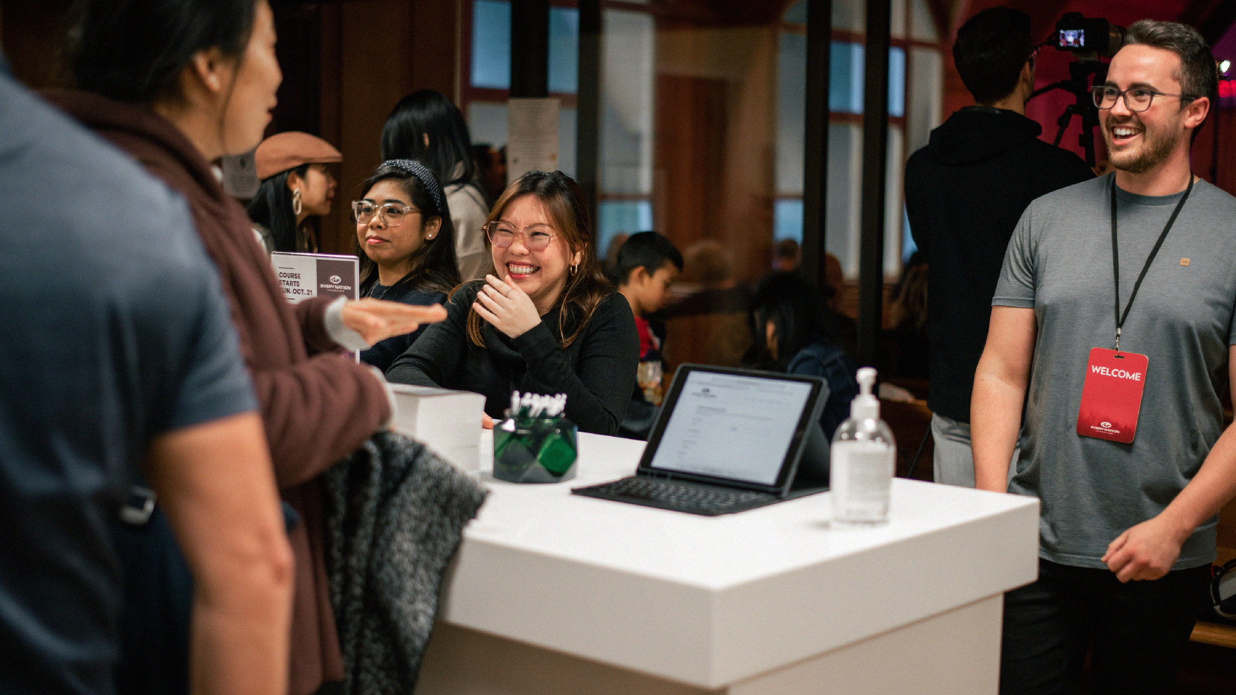People at a reception desk smiling and talking in a busy indoor event with a woman wearing glasses and a black top, a man with a lanyard that says 'WELCOME,' and other attendees in the background.