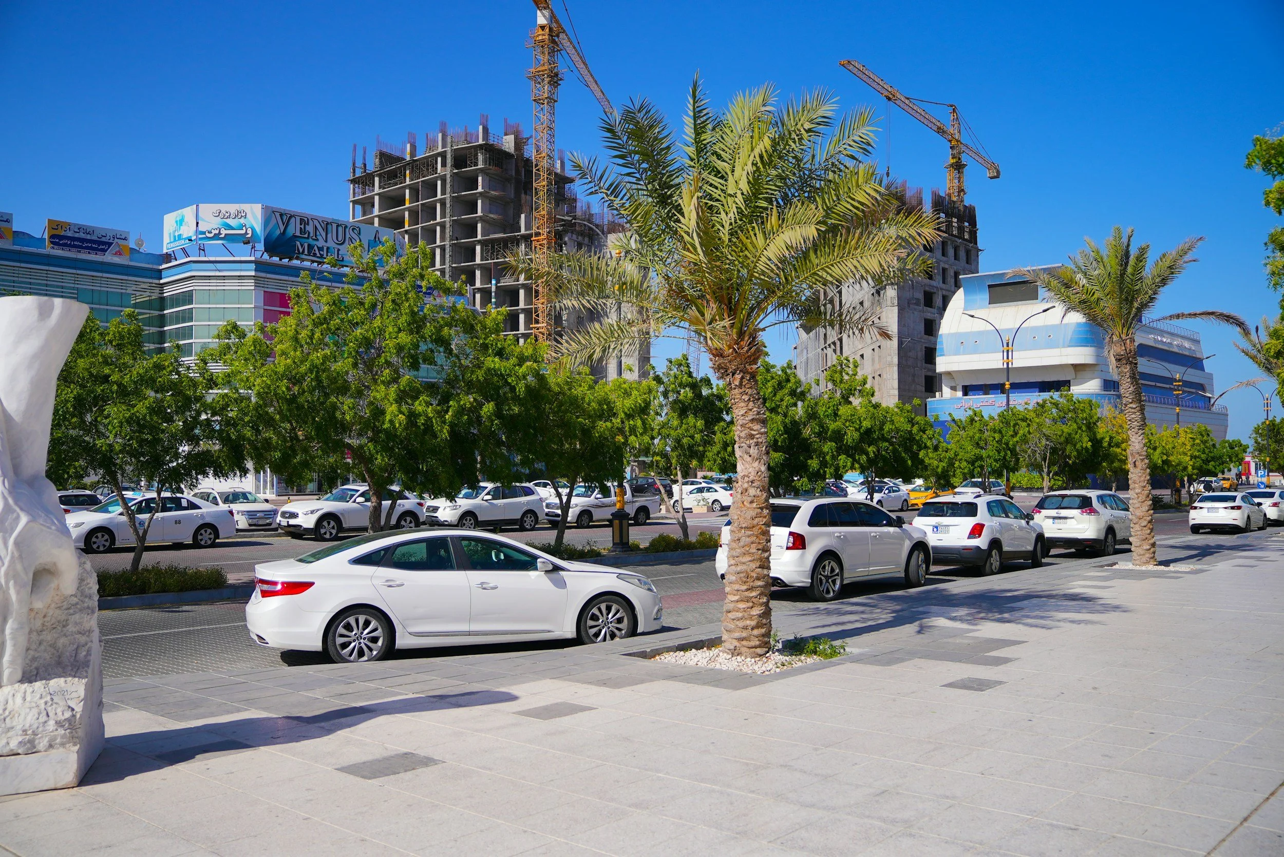 Street view with parked cars, palm trees, a building under construction, and modern buildings in the background, under a clear blue sky.