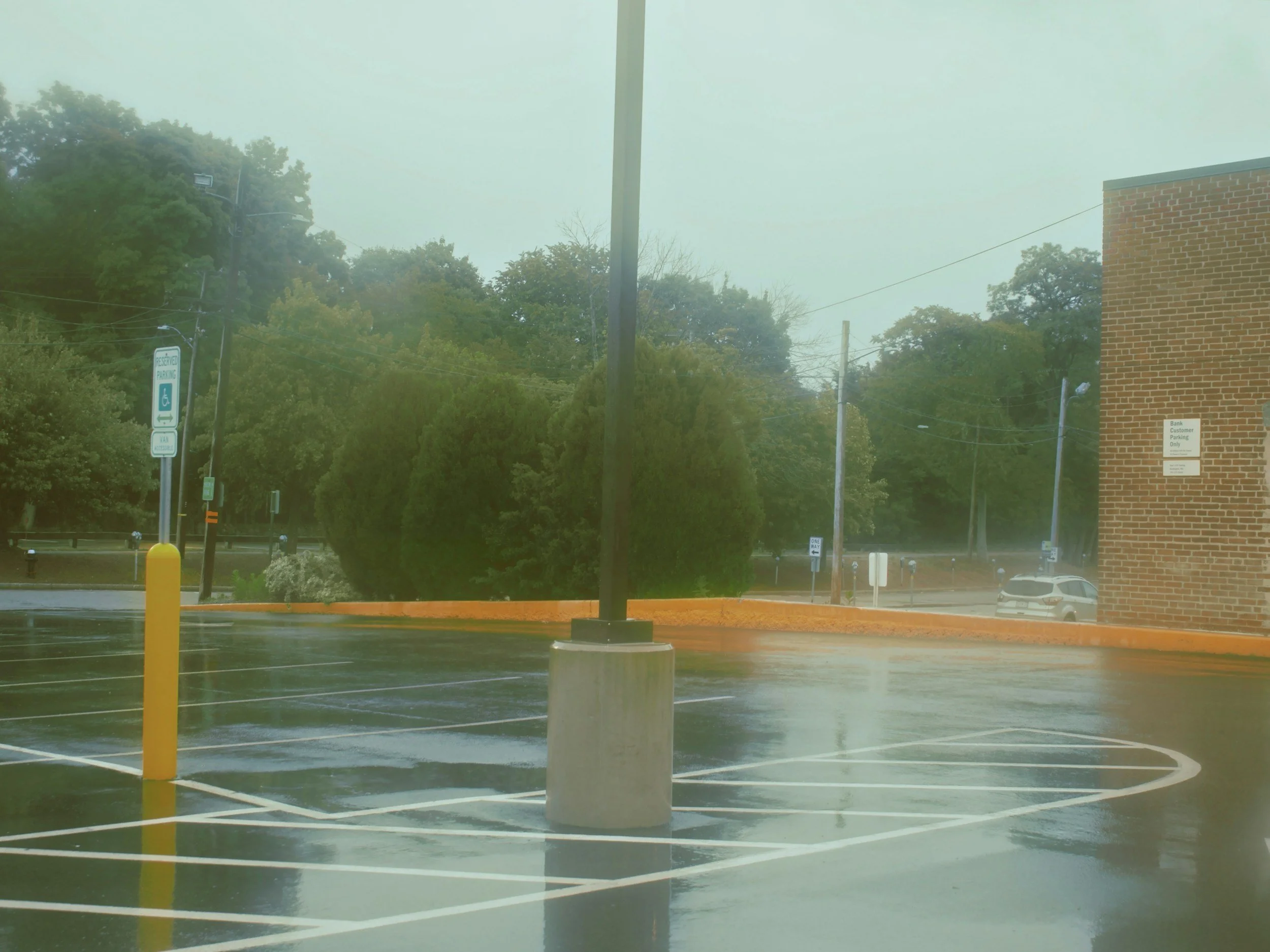 Empty parking lot with wet surface, yellow safety post, brick building corner, and greenery in background on a rainy day.