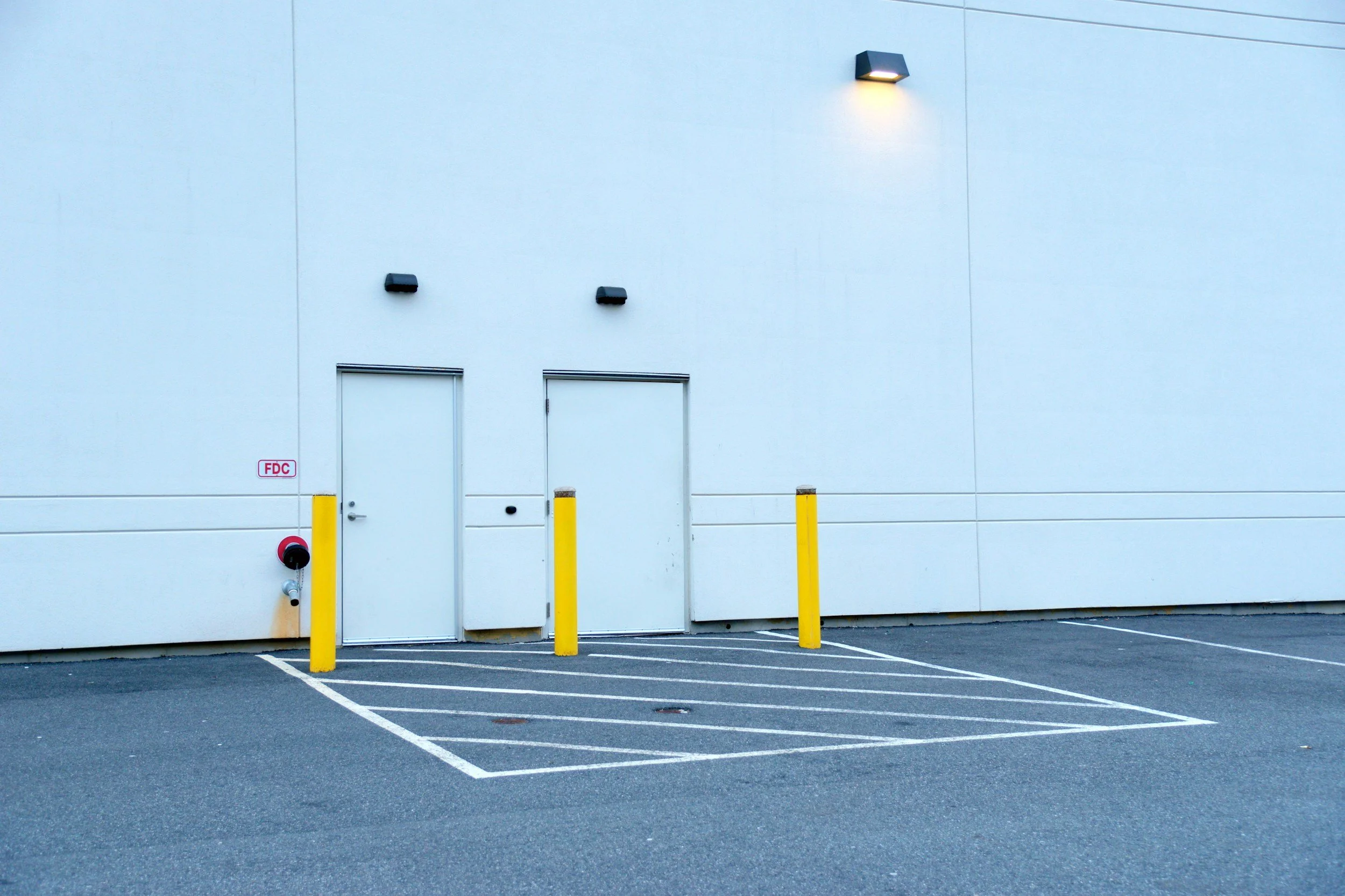 Empty parking lot with two yellow safety bollards, a white wall with two doors, a fire hydrant, and exterior lighting.