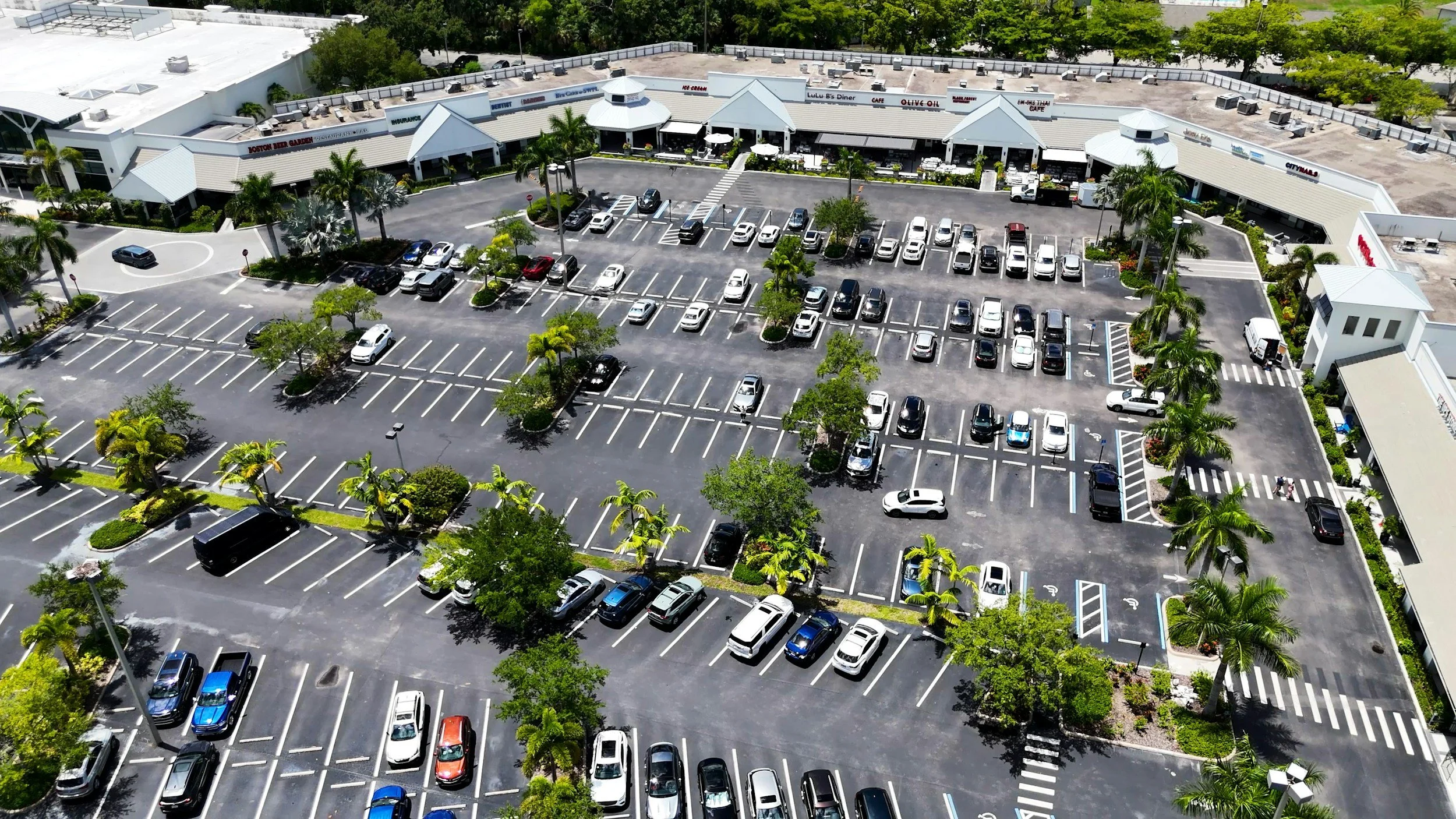 Aerial view of a shopping center parking lot with cars parked in marked spaces, landscaped with trees and bushes, adjacent to storefronts with signs, including a cafe and retail stores, with clear weather and some building rooftops visible.