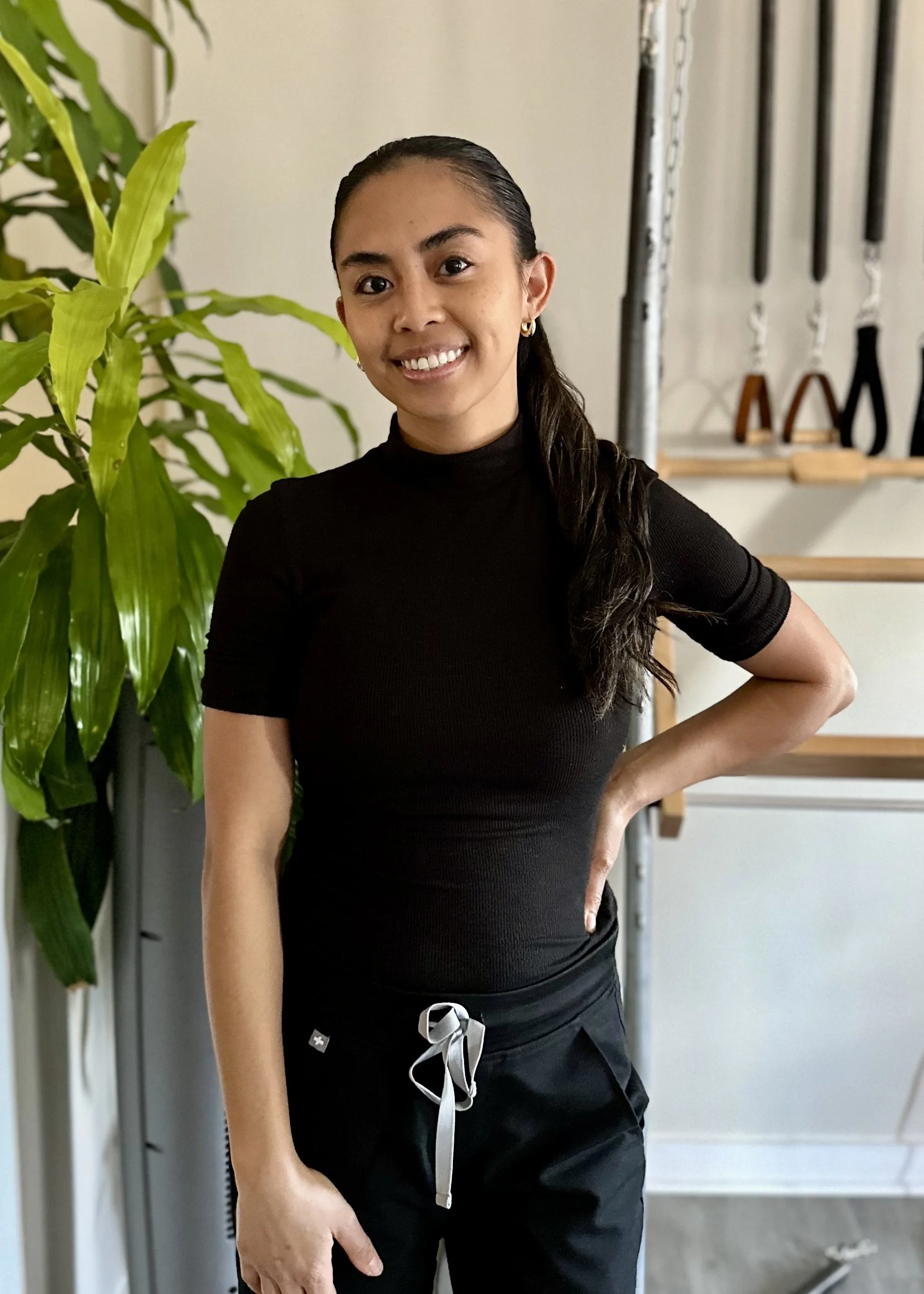 A woman in a black turtleneck and black pants standing indoors with a green plant to her left and gym equipment in the background.