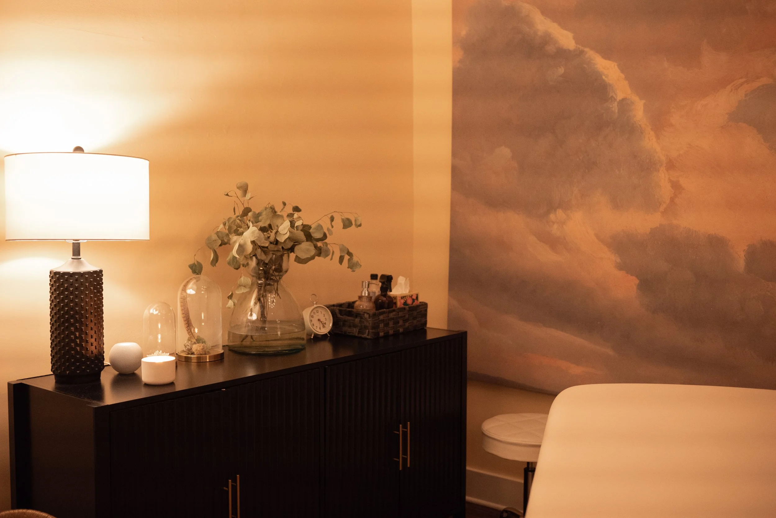 A dark wooden sideboard with various decorative items including a large lamp, a vase with dried flowers, a small clock, and other small objects. On the wall behind is a large cloud-themed mural with pink and orange hues, creating a warm ambiance.